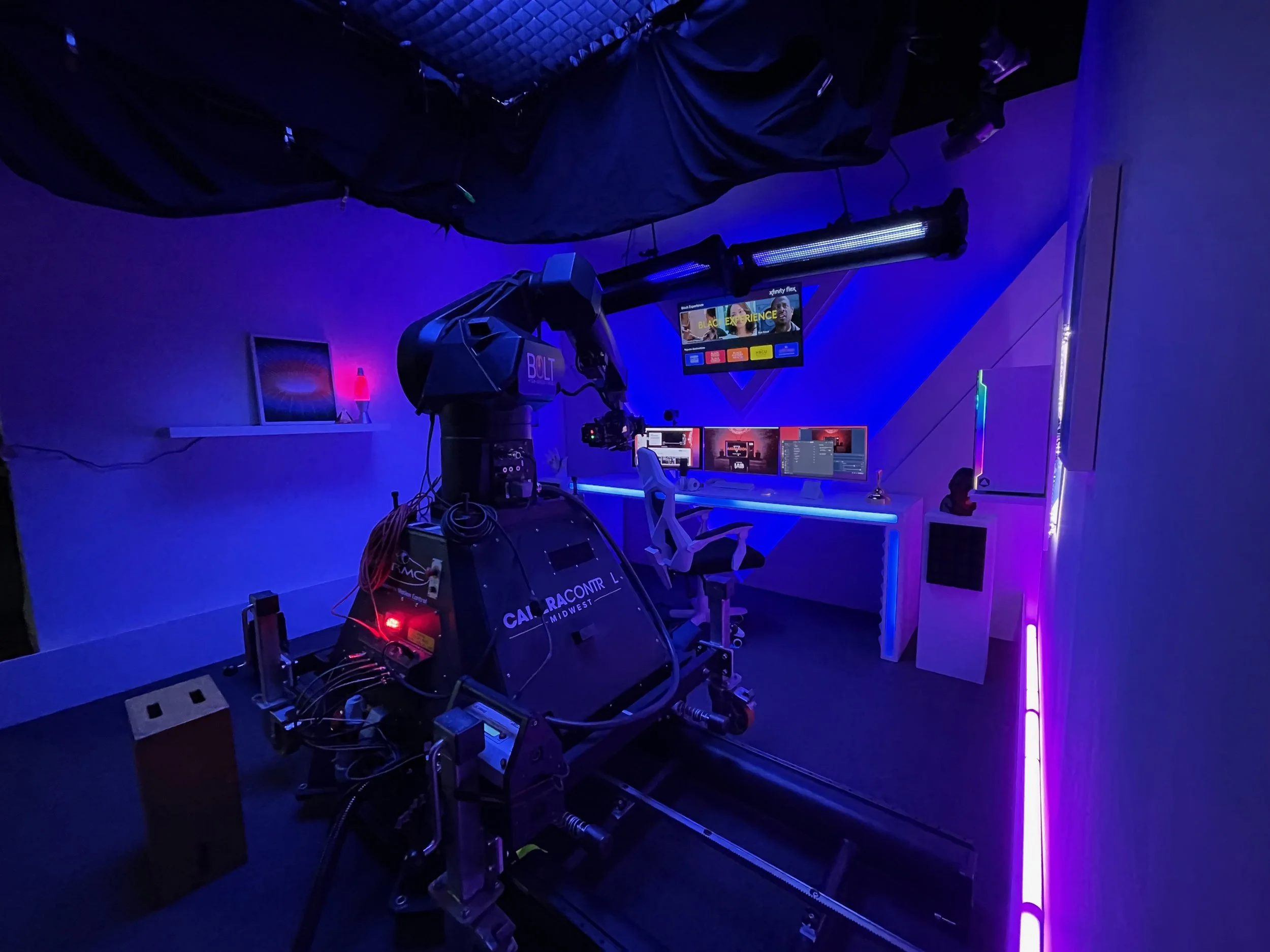 A room with various technological equipment including computer monitors, a robot arm labeled 'BOLT', and a television screen displaying virtual content. The room has blue and purple lighting with a professional or experimental atmosphere.