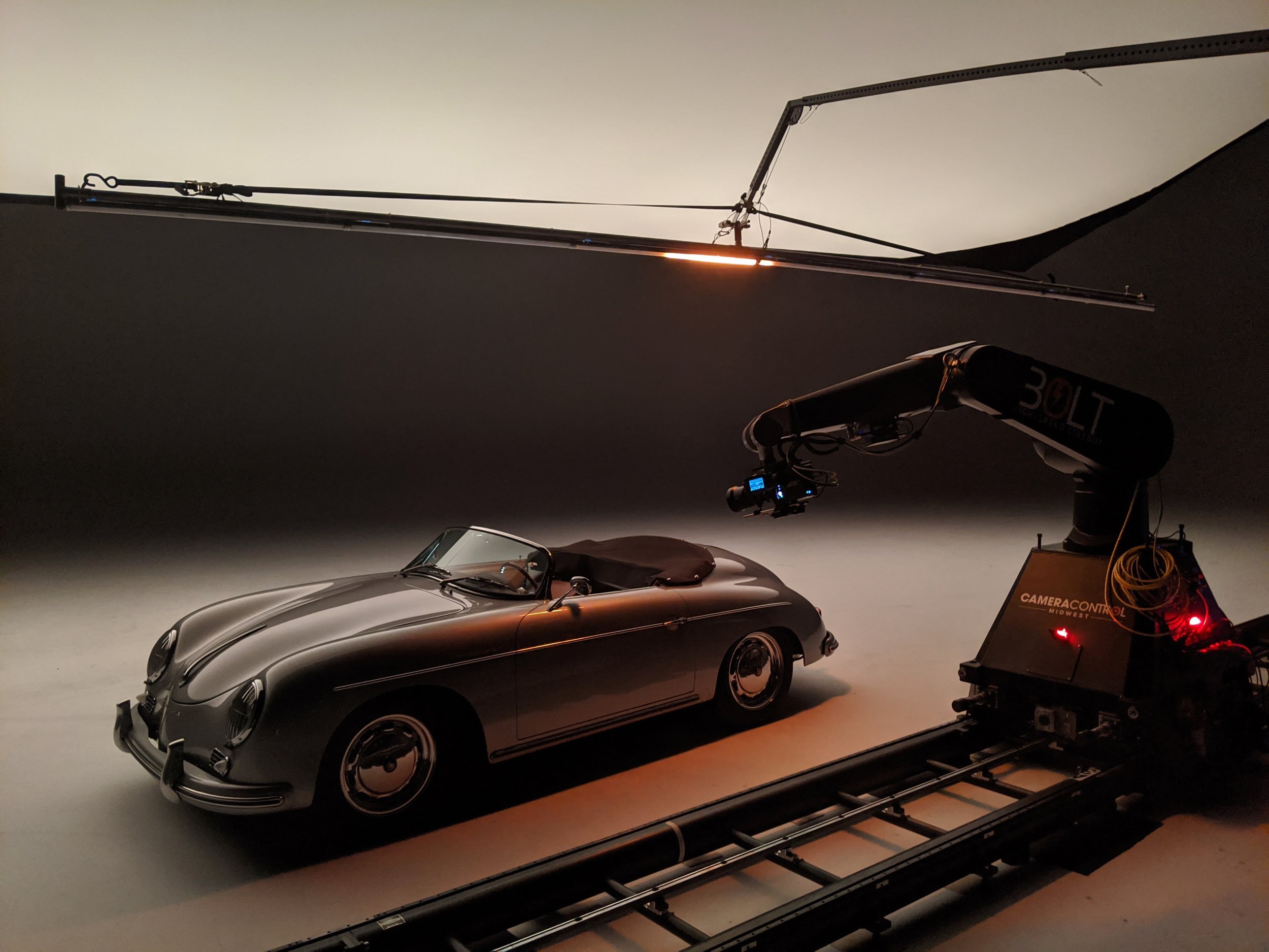 A silver vintage convertible sports car positioned on a rotating platform with a racing helmet on the seat, being filmed with a camera mounted on a track, and large studio lights overhead.