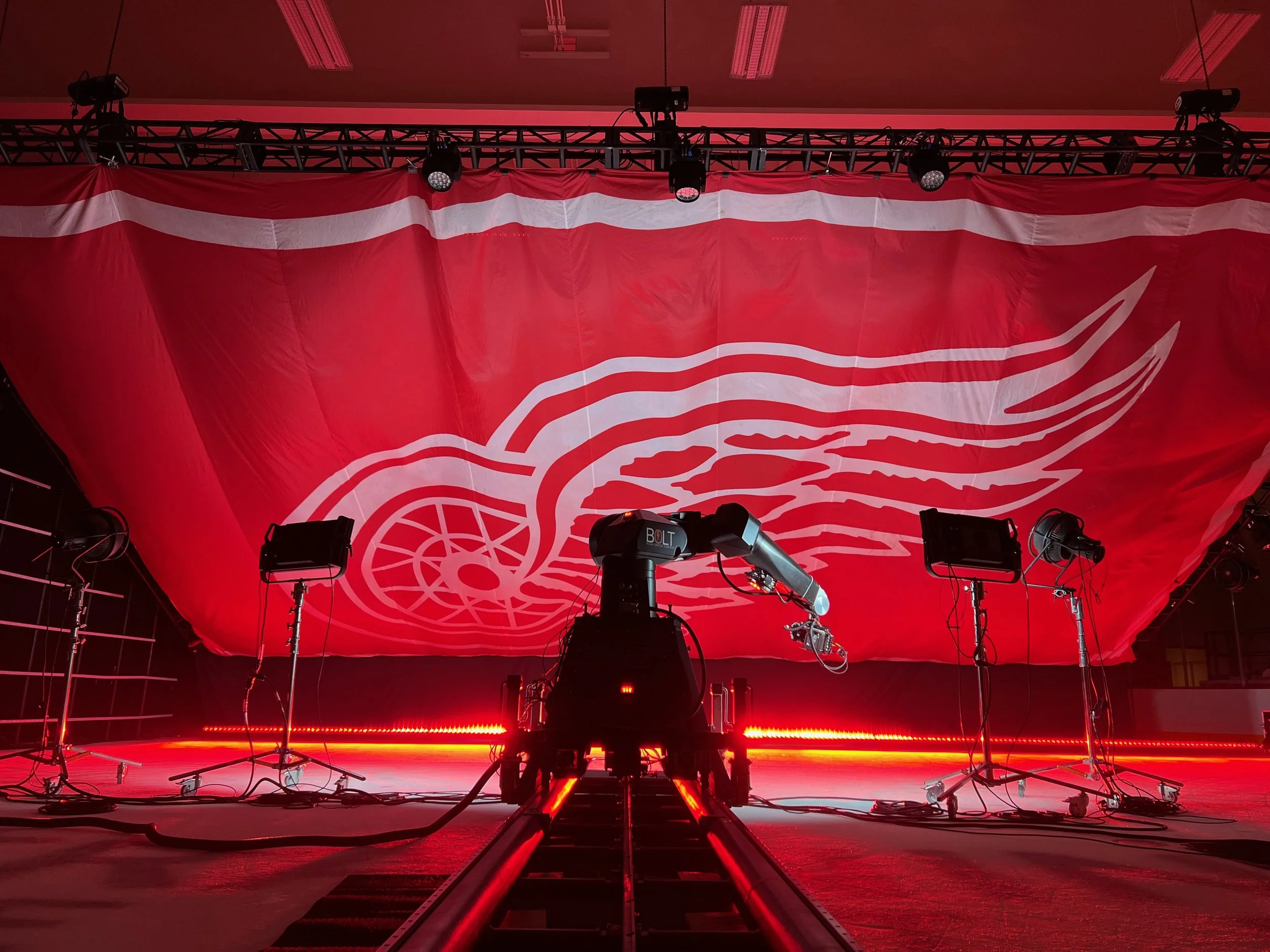 Stage with red lighting, large red and white Detroit Red Wings logo banner, stage equipment, and lighting rigs.