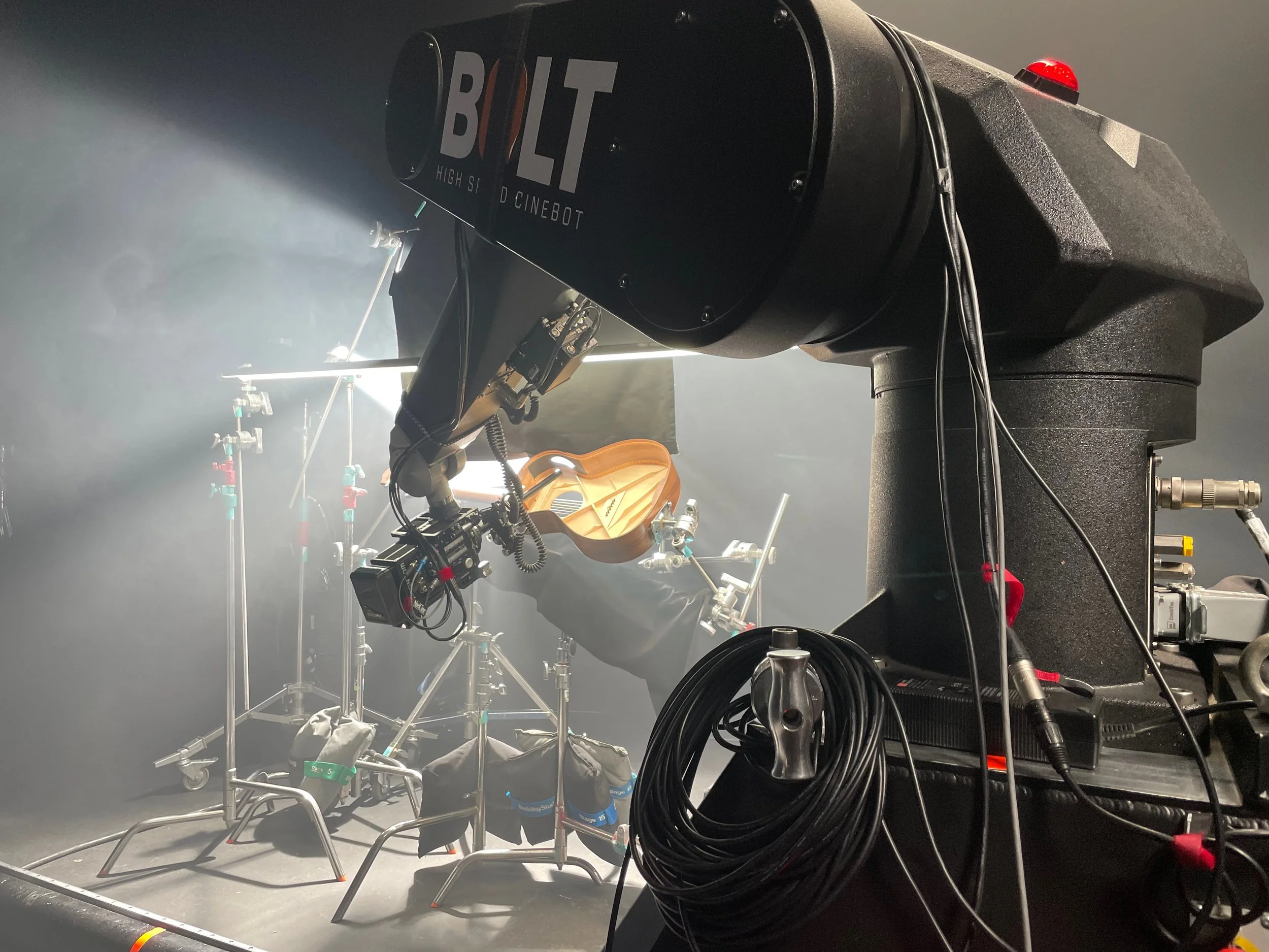 Close-up of a large black camera or projector with a red button on top and papers or labels attached to its side, in a room with studio lighting equipment and a musical instrument in the background.