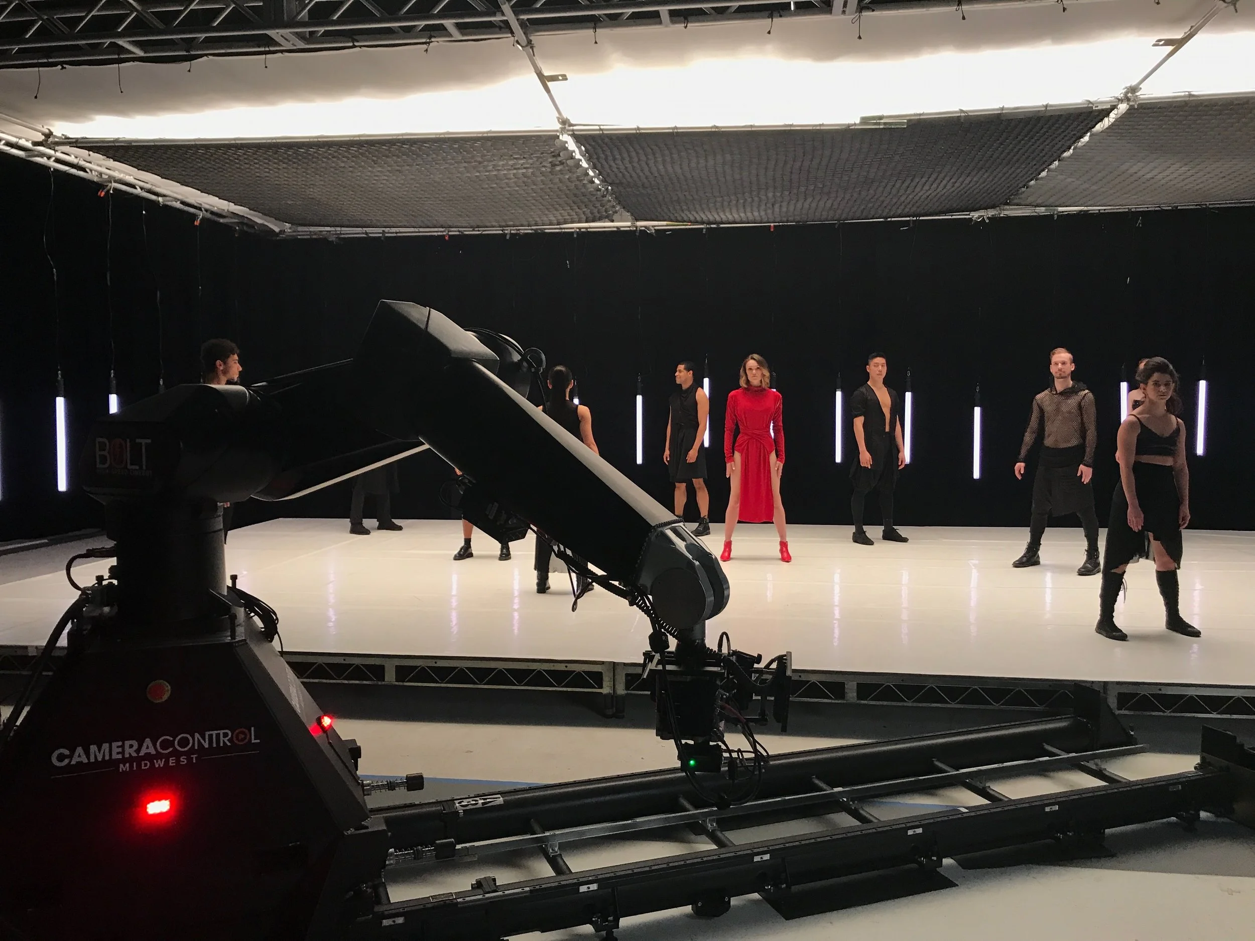 A photography studio with six models standing on a black backdrop, illuminated by vertical light tubes, with professional cameras and equipment in the foreground.