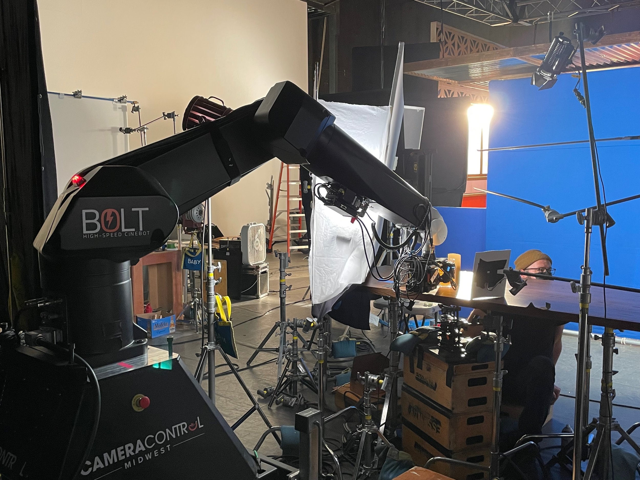 A behind-the-scenes view of a film or photography studio. It includes professional lighting equipment, a camera labeled 'Bolt High-Speed Cinebot,' a blue screen on the right, and various accessories and tools scattered around. There is a person visib