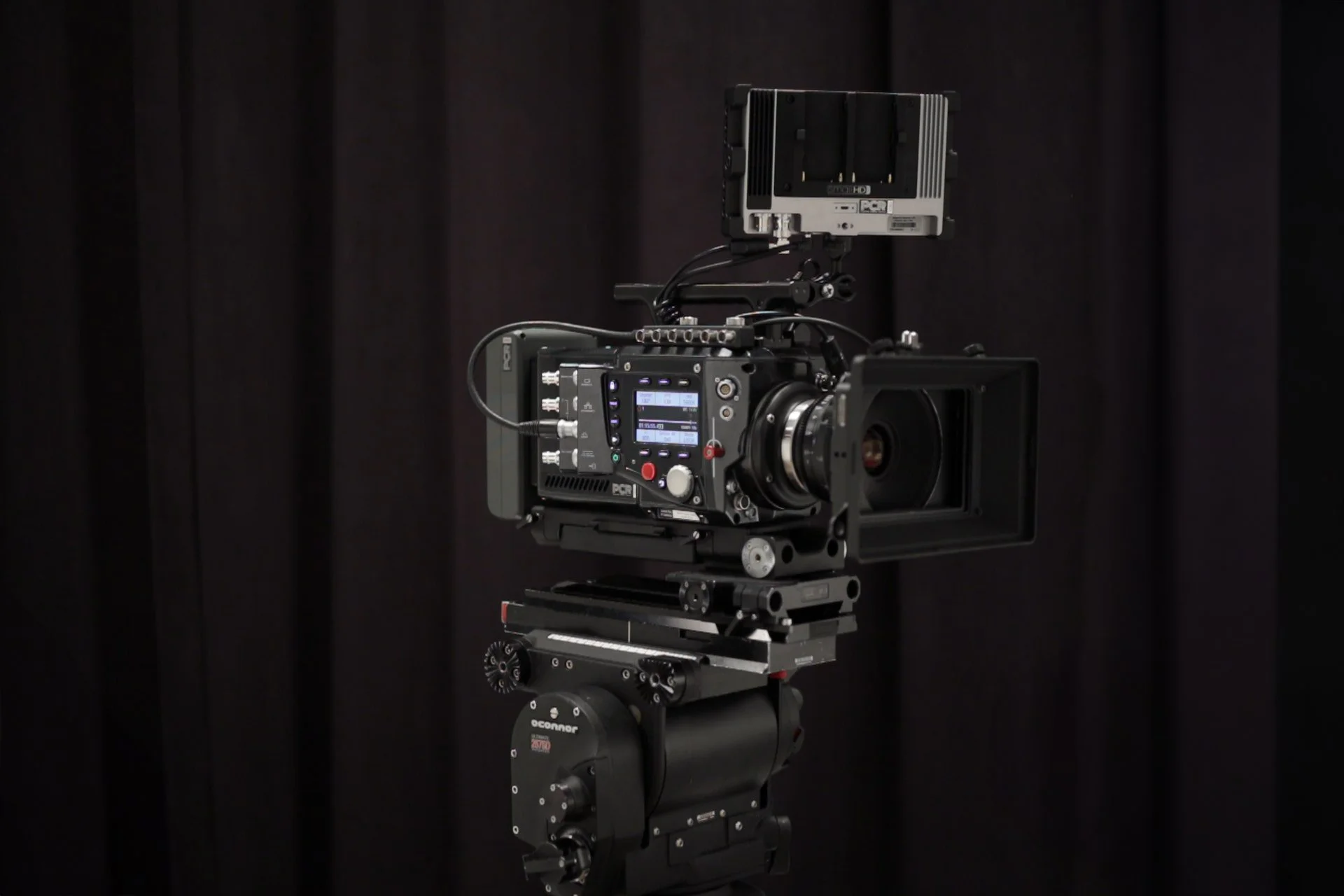 Professional cinema camera mounted on a tripod with a matte box and external monitor, set against a dark curtain background.