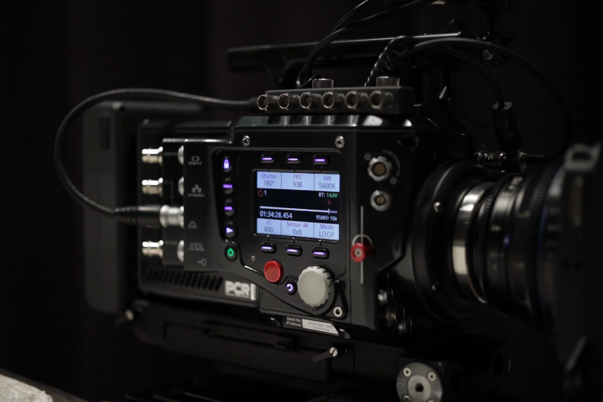 Close-up of a professional digital cinema camera with adjustable settings on a small display screen, surrounded by various buttons, knobs, and cables against a dark background.