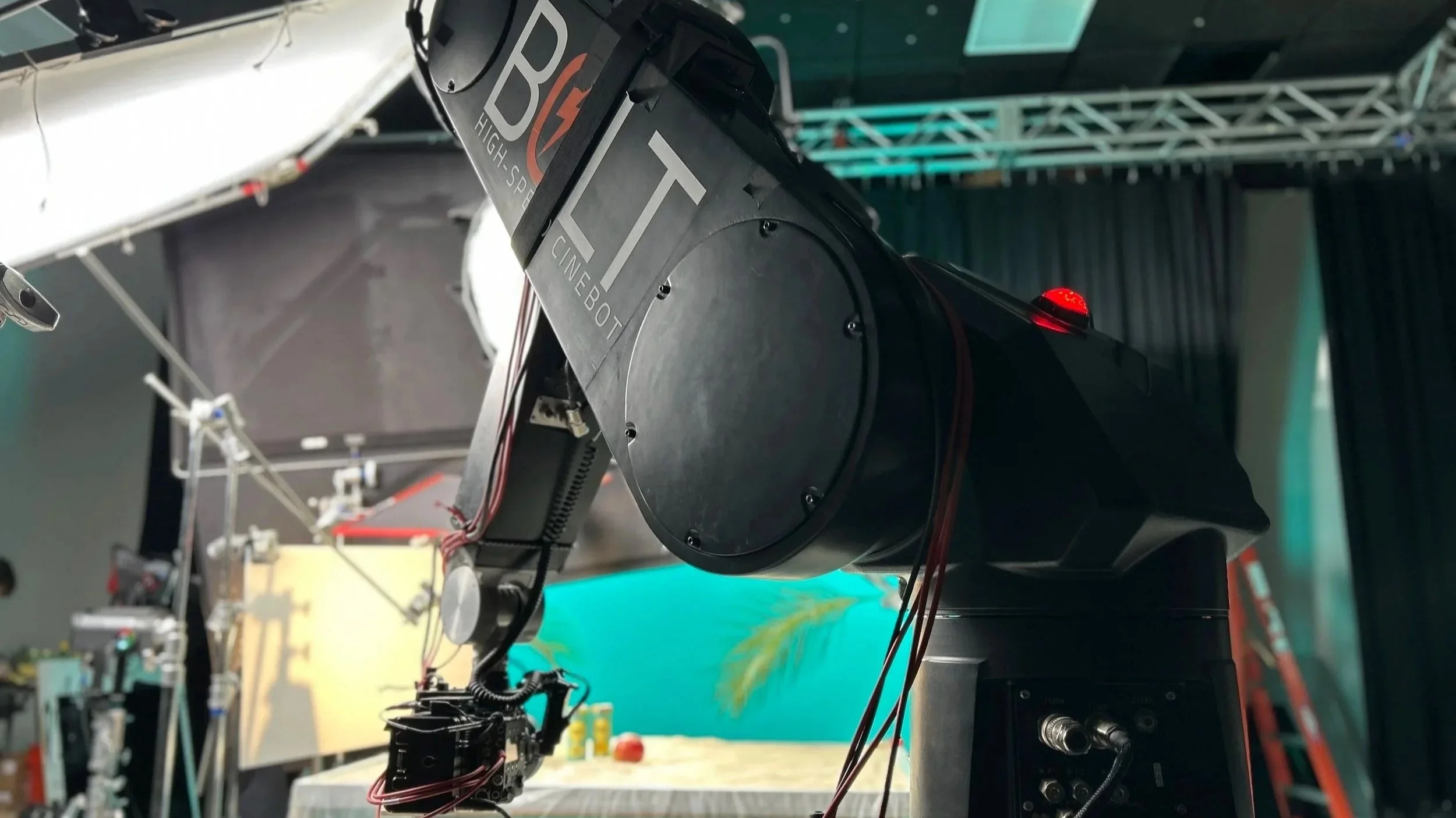 A robotic arm with a camera or sensor attached, labeled 'BOLT Cineboot,' appears to be part of a robotics or automation project in a studio setting. The background shows equipment and a colorful backdrop.