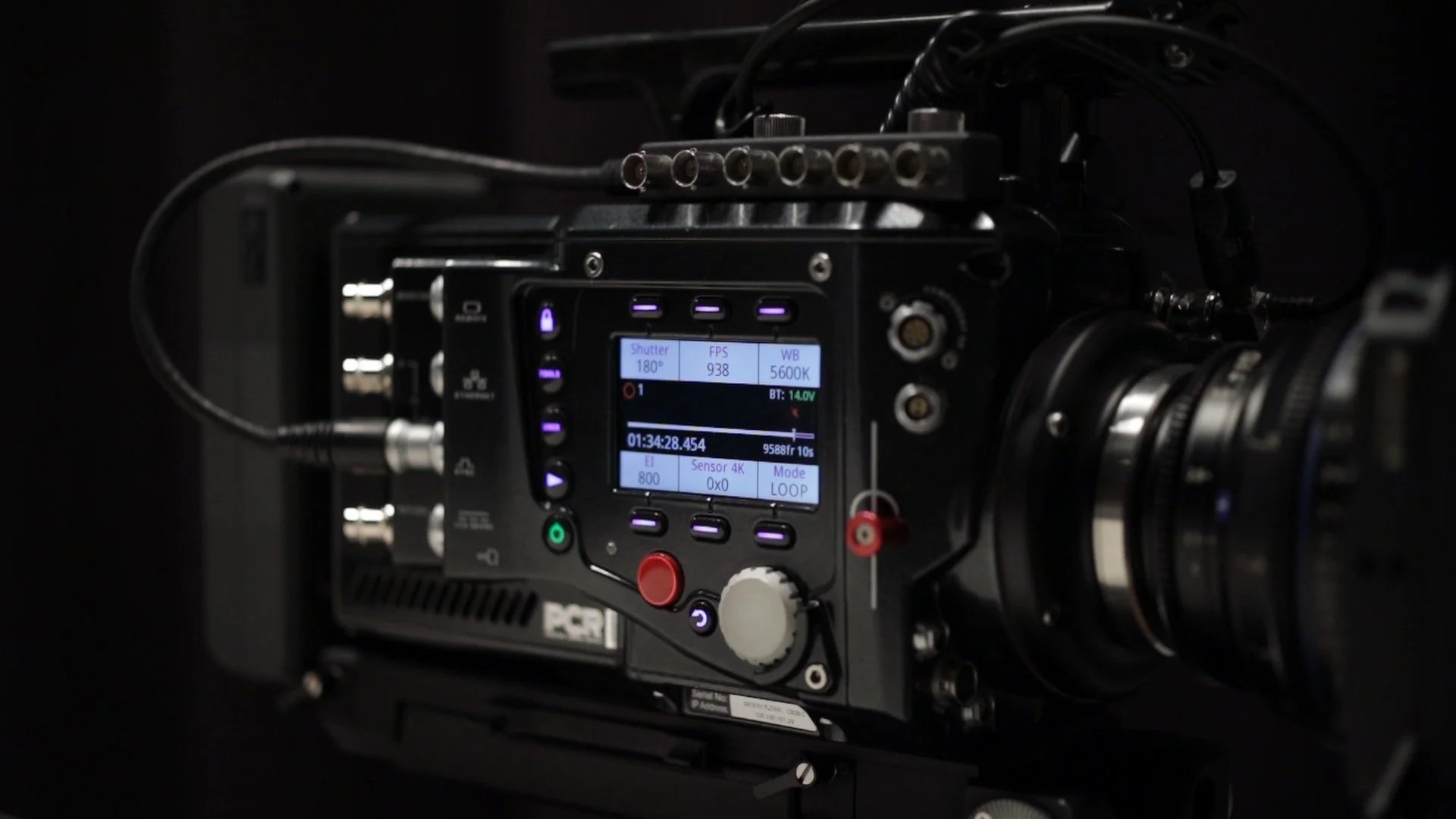 Close-up of professional video camera with digital screen displaying settings, surrounded by black equipment and cables in a dark environment.