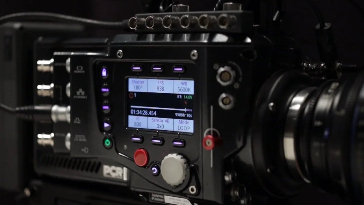 Close-up of a professional video camera with an electronic control panel displaying settings, attached to a large lens.