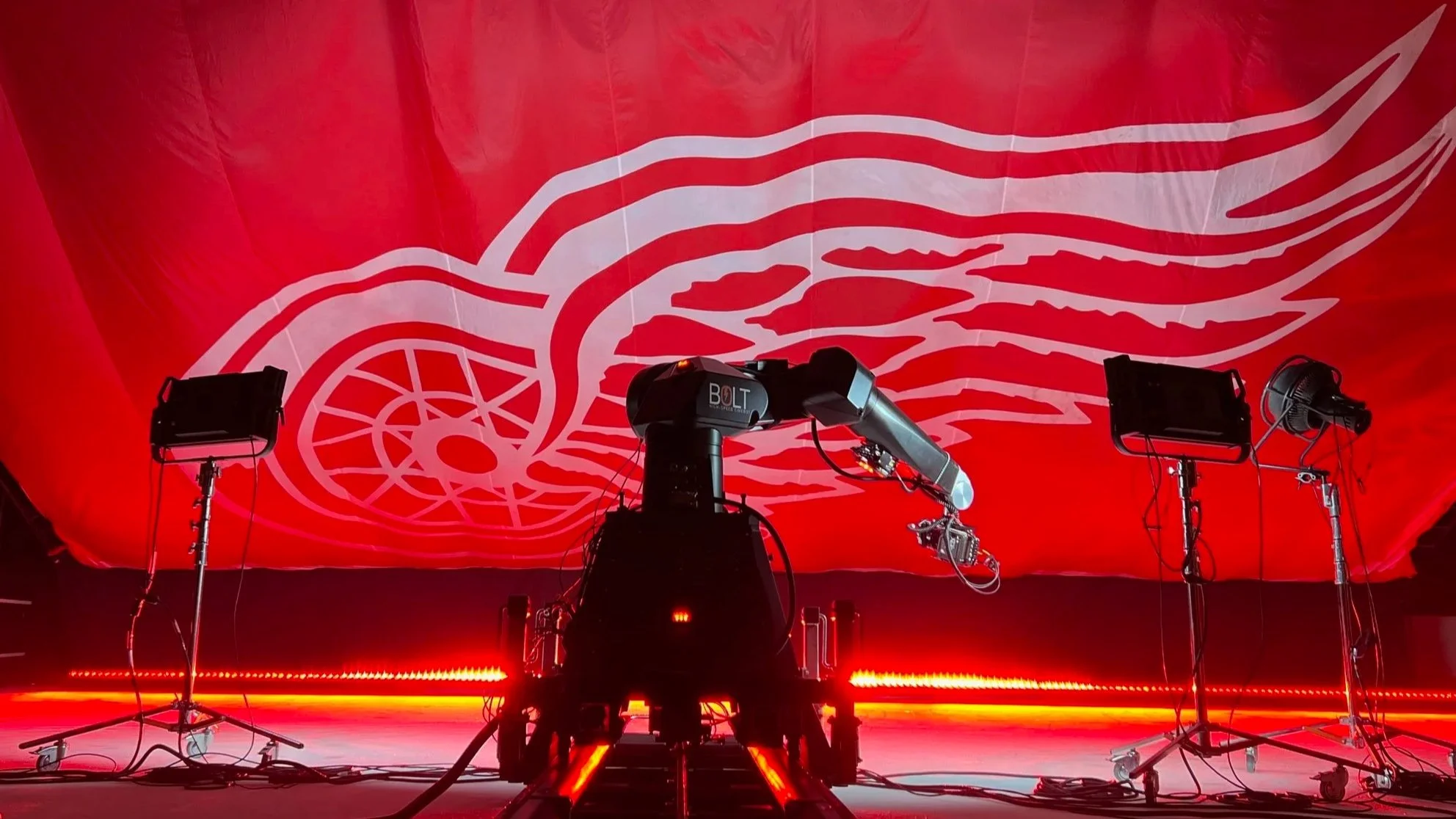 Stage setup with lighting equipment and a large red backdrop featuring a stylized white Detroit Red Wings logo, including a wing and wheel design.