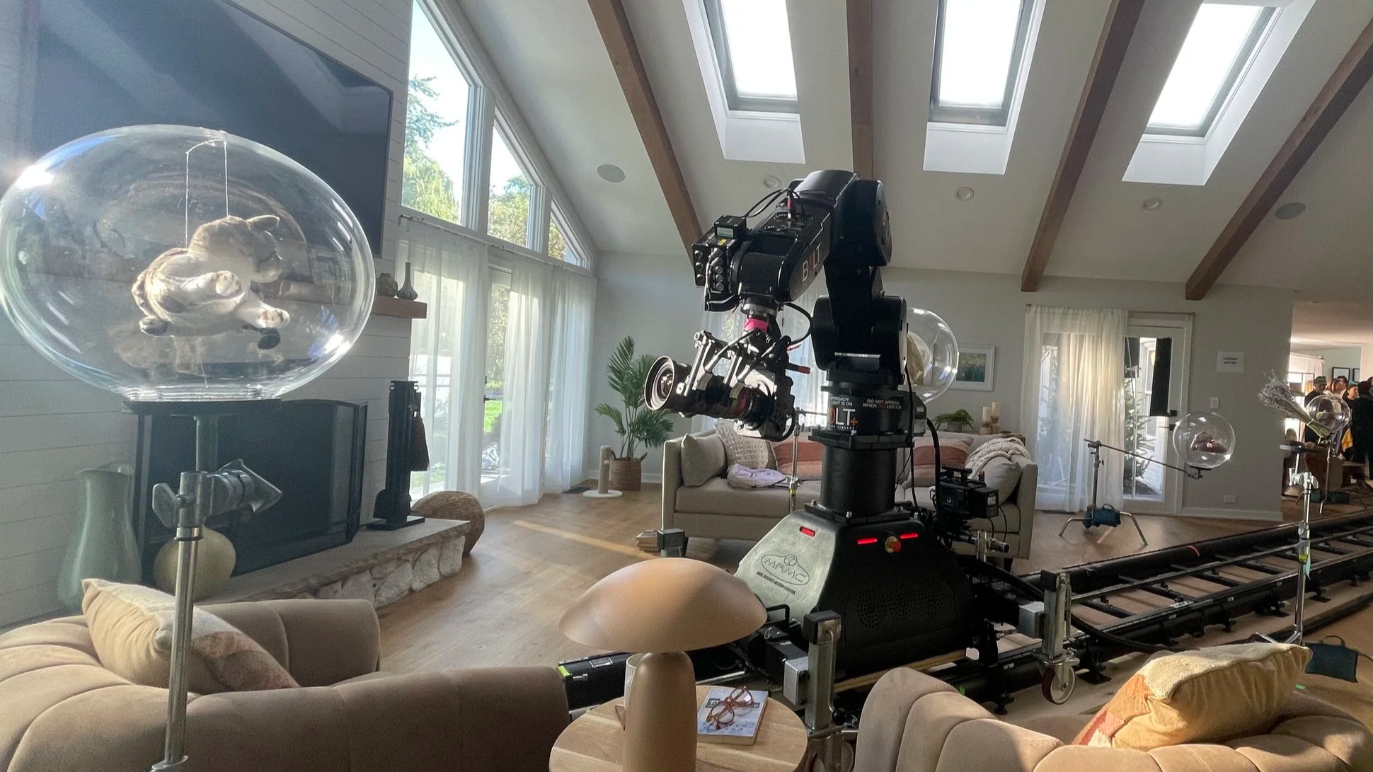 Interior of a well-lit living room with large windows, a ceiling with skylights, and a camera setup on a dolly for filming. The room has a beige sofa, a brick fireplace, and various decorative items, with several people in the background near a gathering area.