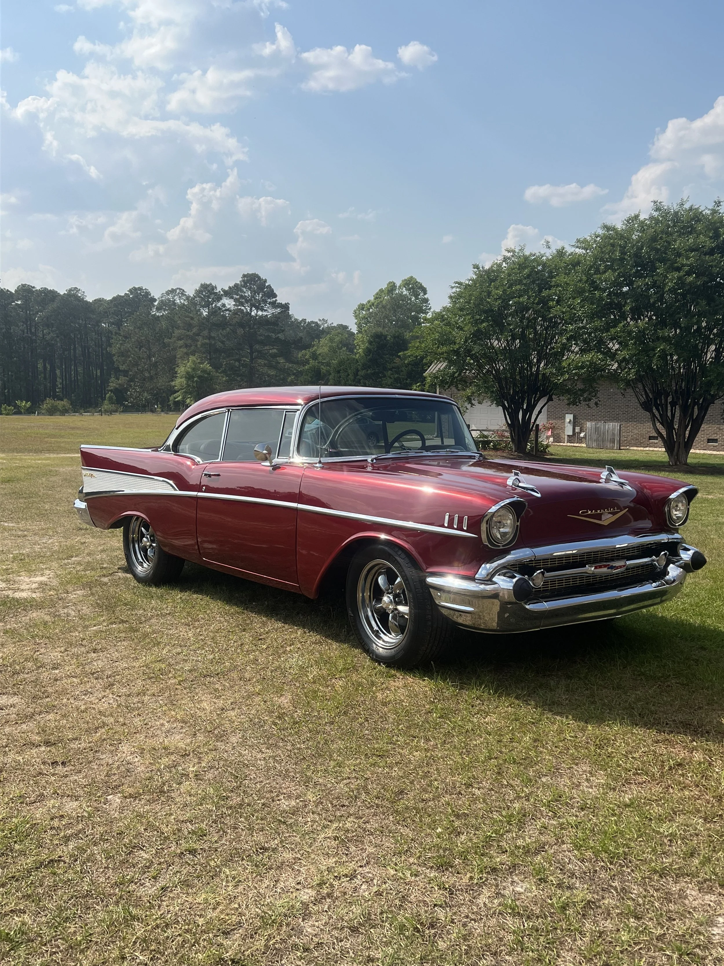 A vintage red and white Chevrolet car parked on a grassy field with trees and a brick house in the background under a partly cloudy sky.