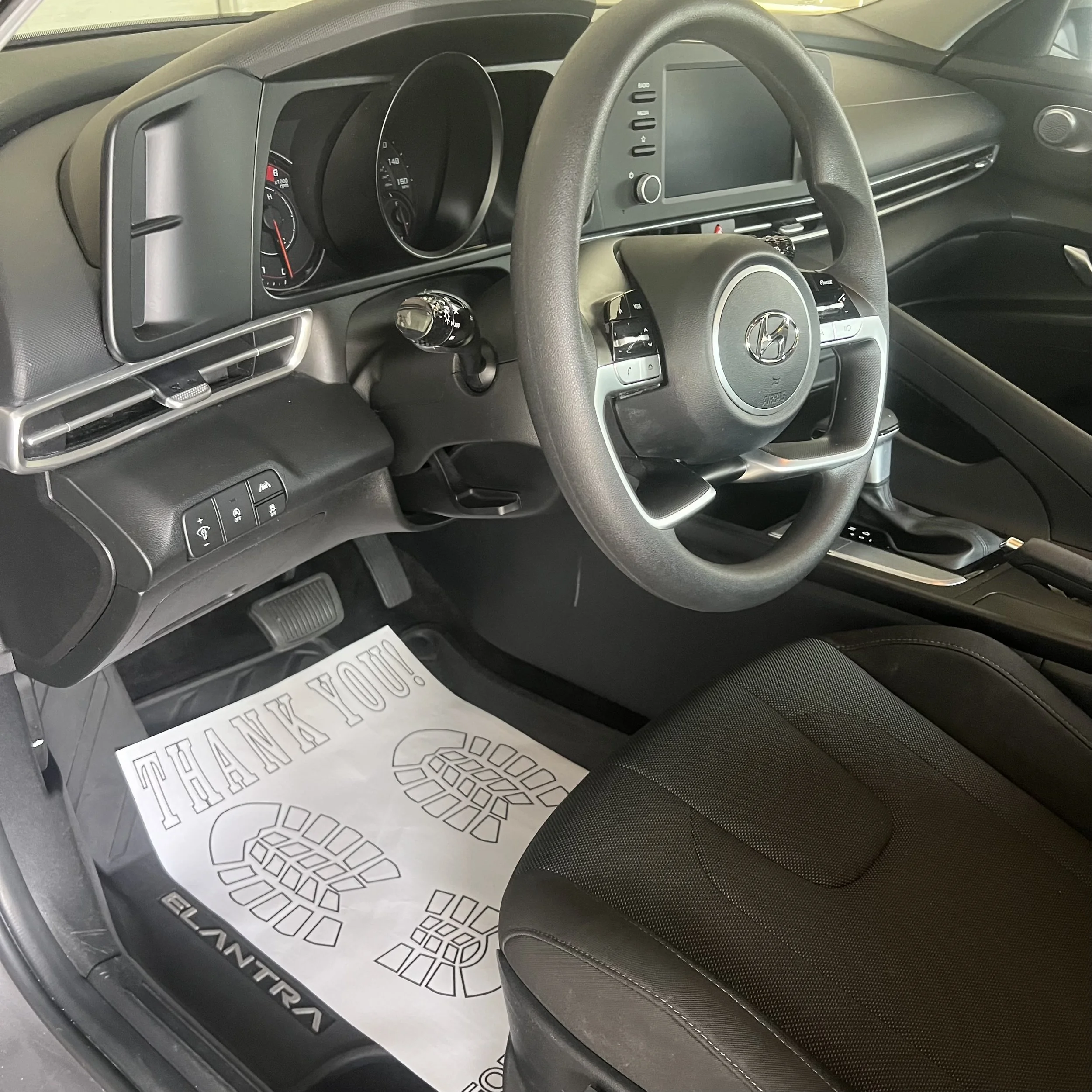 View of the interior of a Hyundai vehicle showing the dashboard, steering wheel, and front seats, with a paper floor mat displaying tire tread pattern on the driver's side.