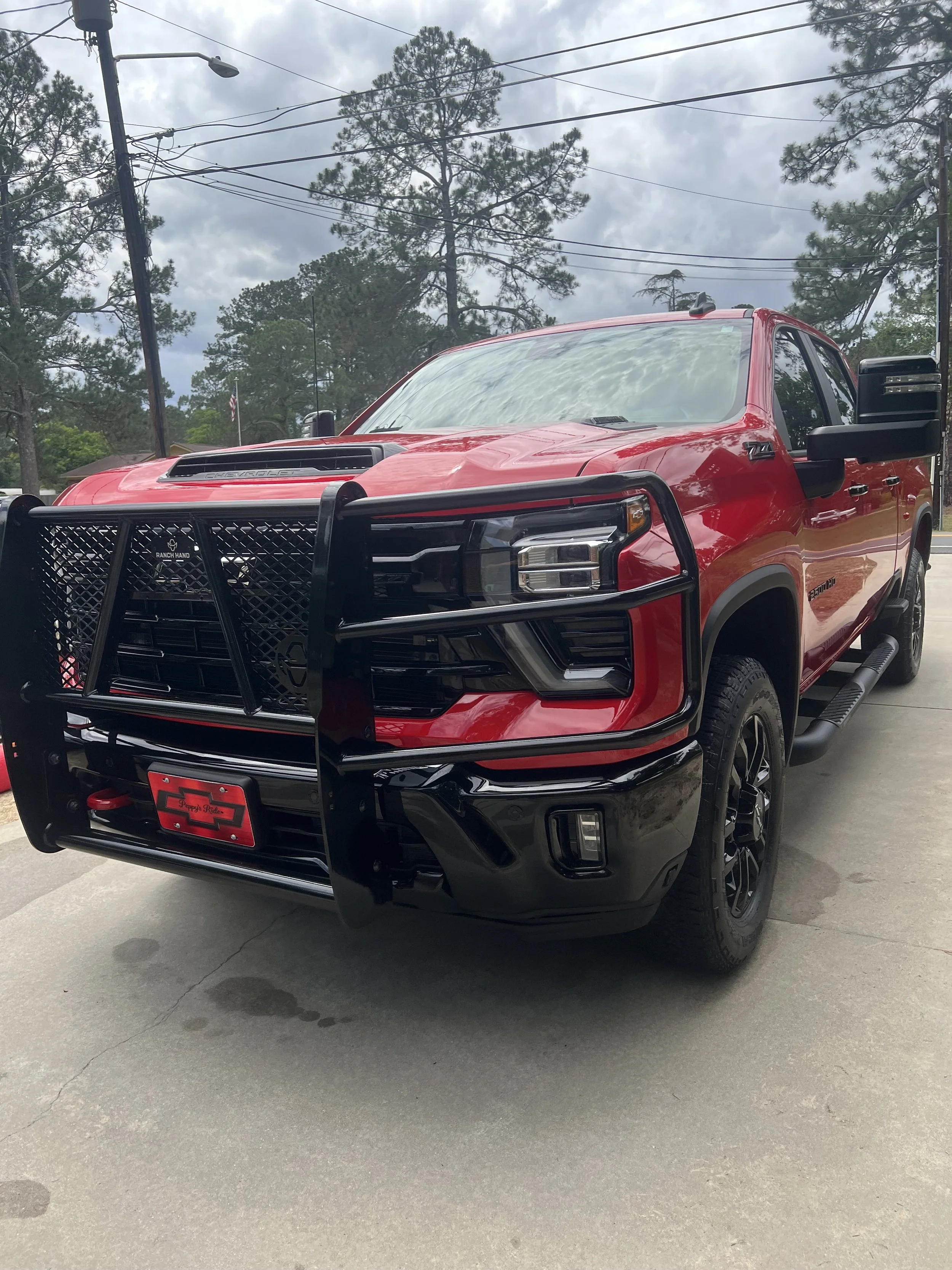 Red pickup truck with a black front grill guard parked on a concrete lot, trees and power lines in the background, overcast sky.