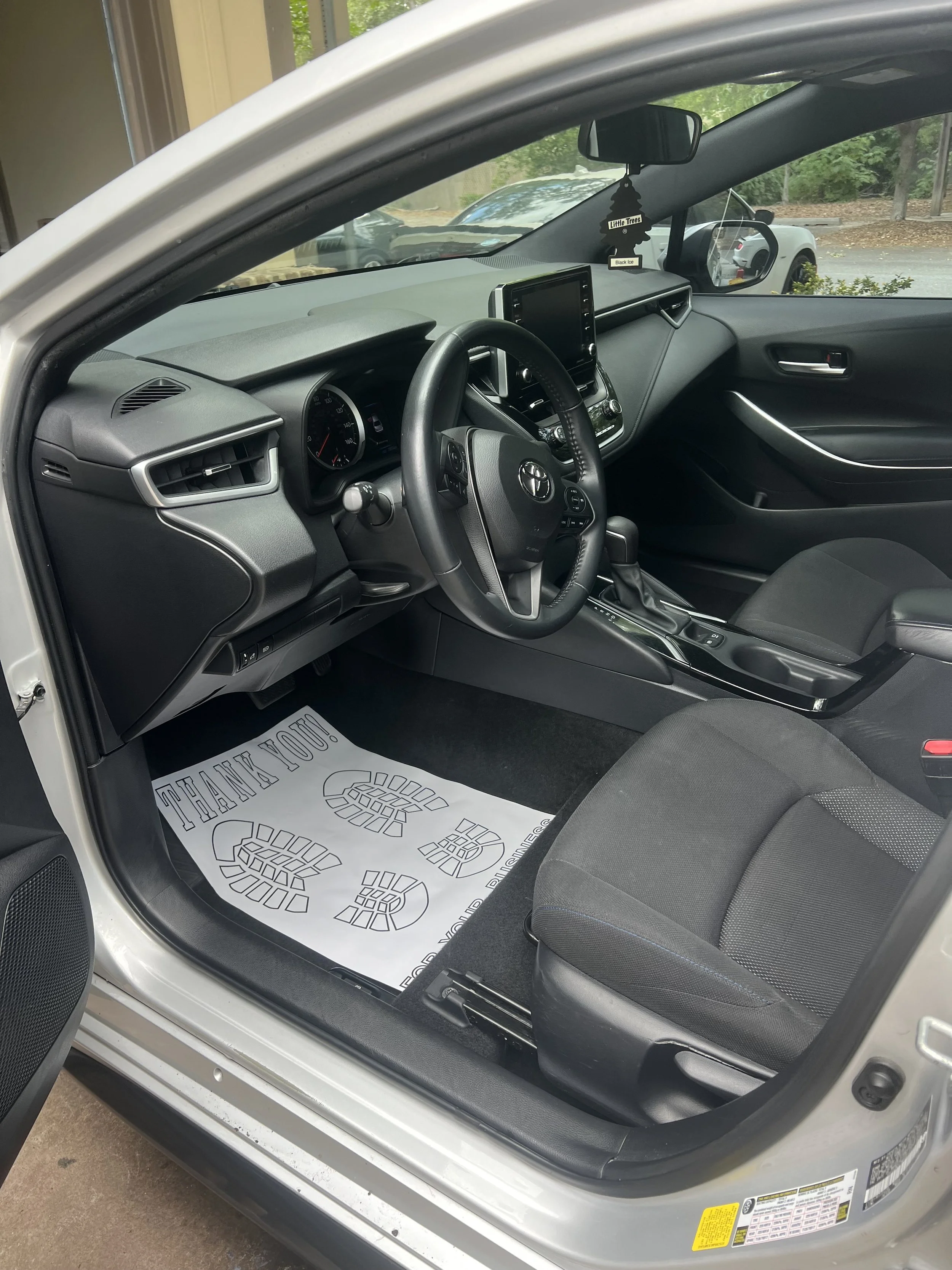 Interior of a car showing the driver's seat, steering wheel, dashboard, and center console. There is a paper floor mat with a message and footprints, and a rearview mirror with hanging air freshener.