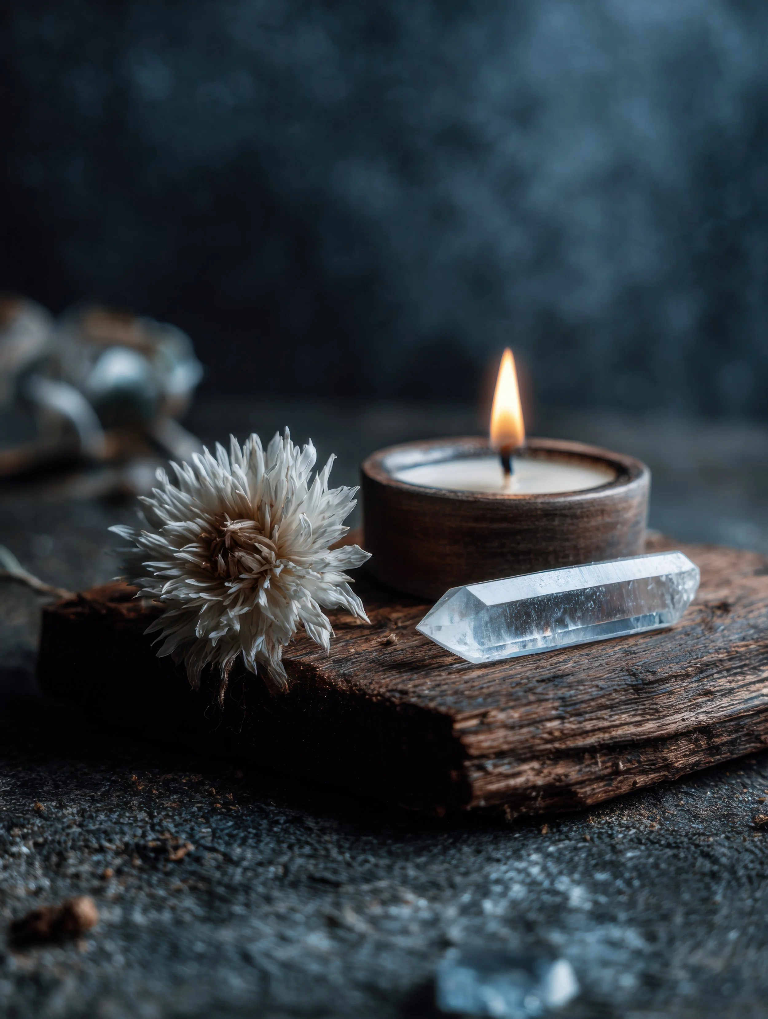 A lit candle in a wooden holder, a clear quartz crystal, and a dried flower on a rustic piece of wood, with a dark, textured background.