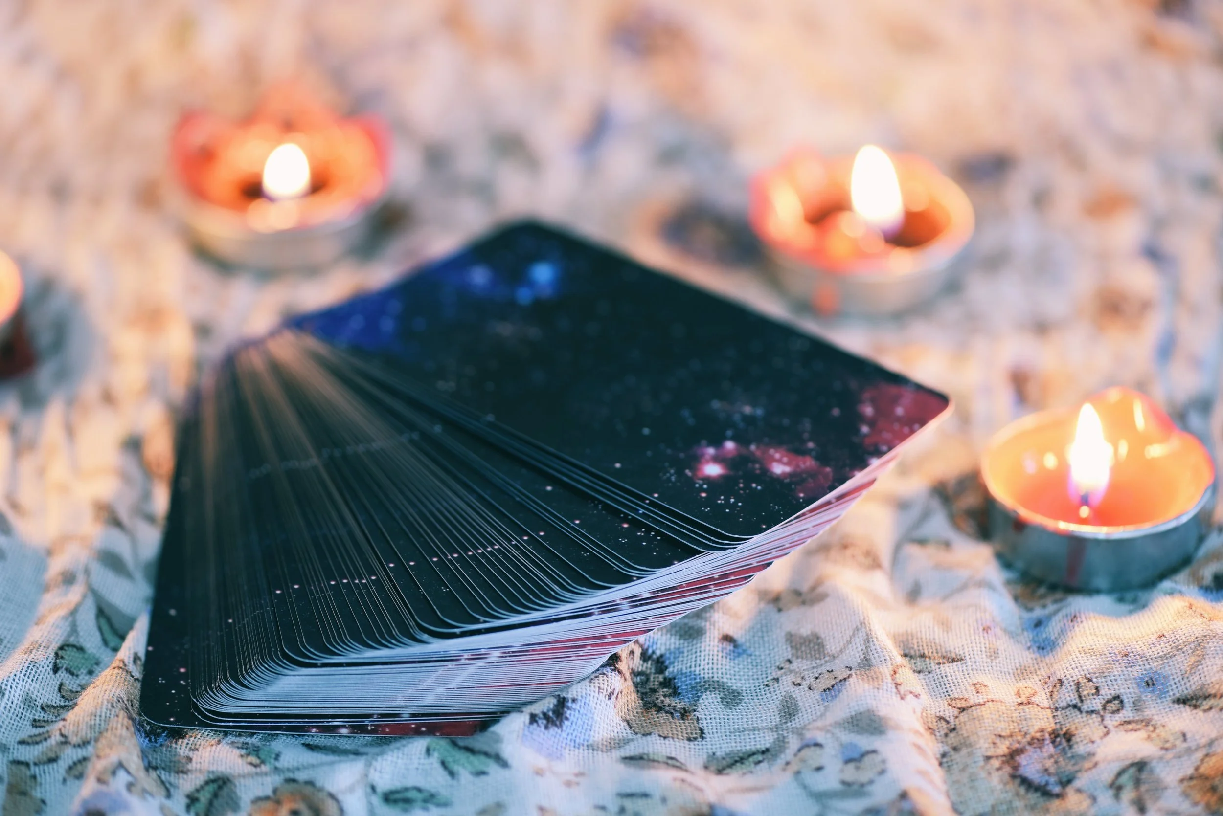A deck of angel cards fanned out on a floral cloth, surrounded by lit candles, with a galaxy-themed design on the cards.
