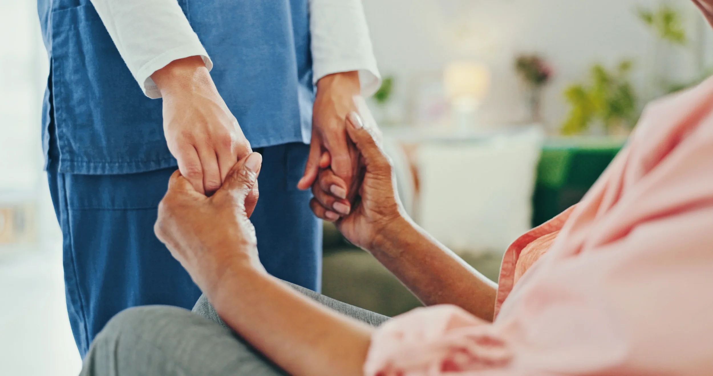Person sitting, holding hands with an elderly woman who is seated. The elderly woman is wearing a peach-colored blouse, and the person is wearing a pink top. The elderly woman appears to be reaching up to the person's hand as they hold hands in a caring gesture. The background is softly blurred, suggesting a warm, indoor setting with plants and soft lighting.