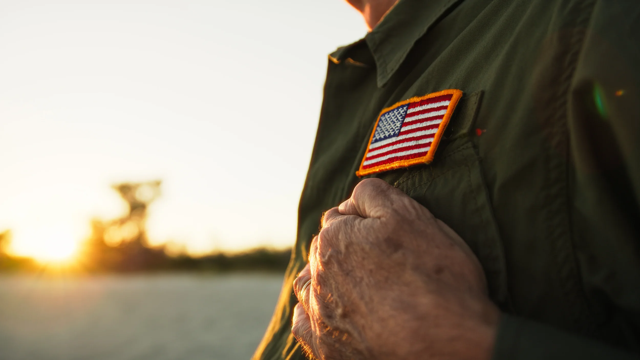 A person wearing a military uniform with a U.S. flag patch on the chest, holding their hand over their heart during sunset.