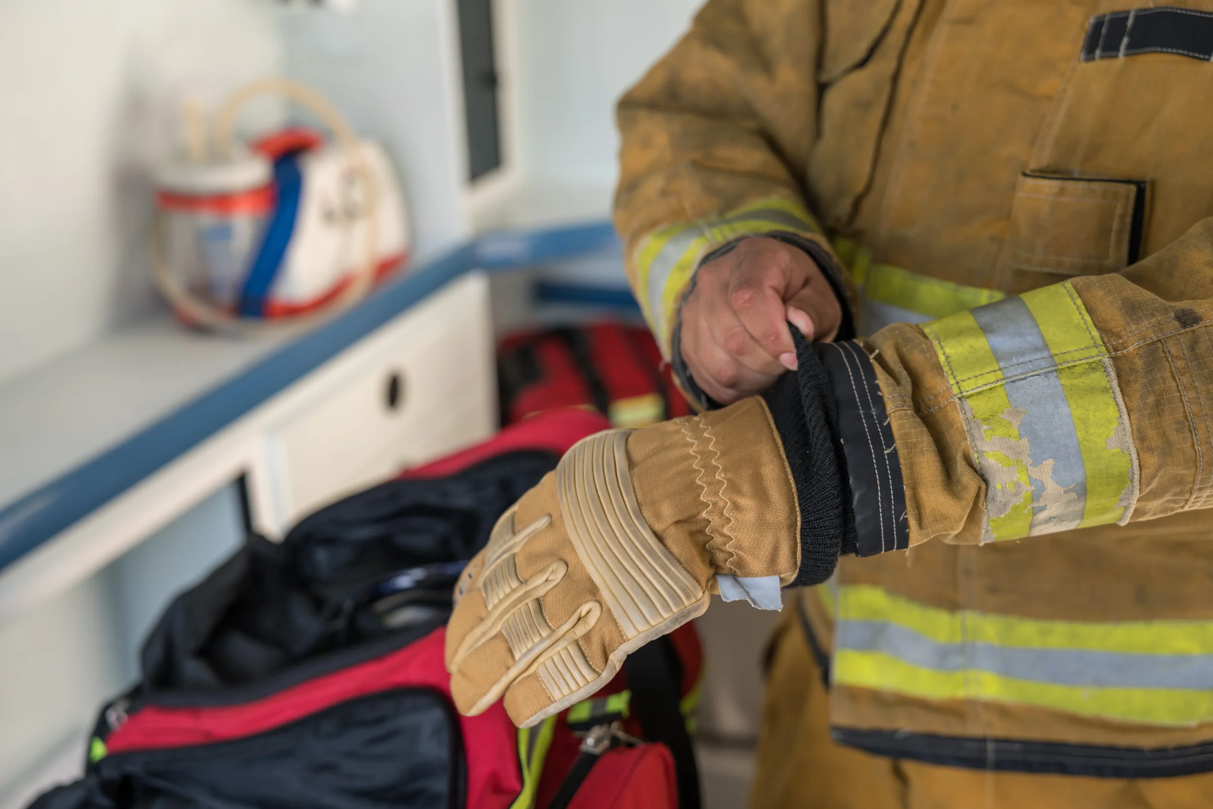 A person dressed in firefighting gear, including a tan fire-resistant suit and gloves, is grabbing the sleeve of another individual wearing a black and red jacket. In the background, fire helmets are visible on a shelf.