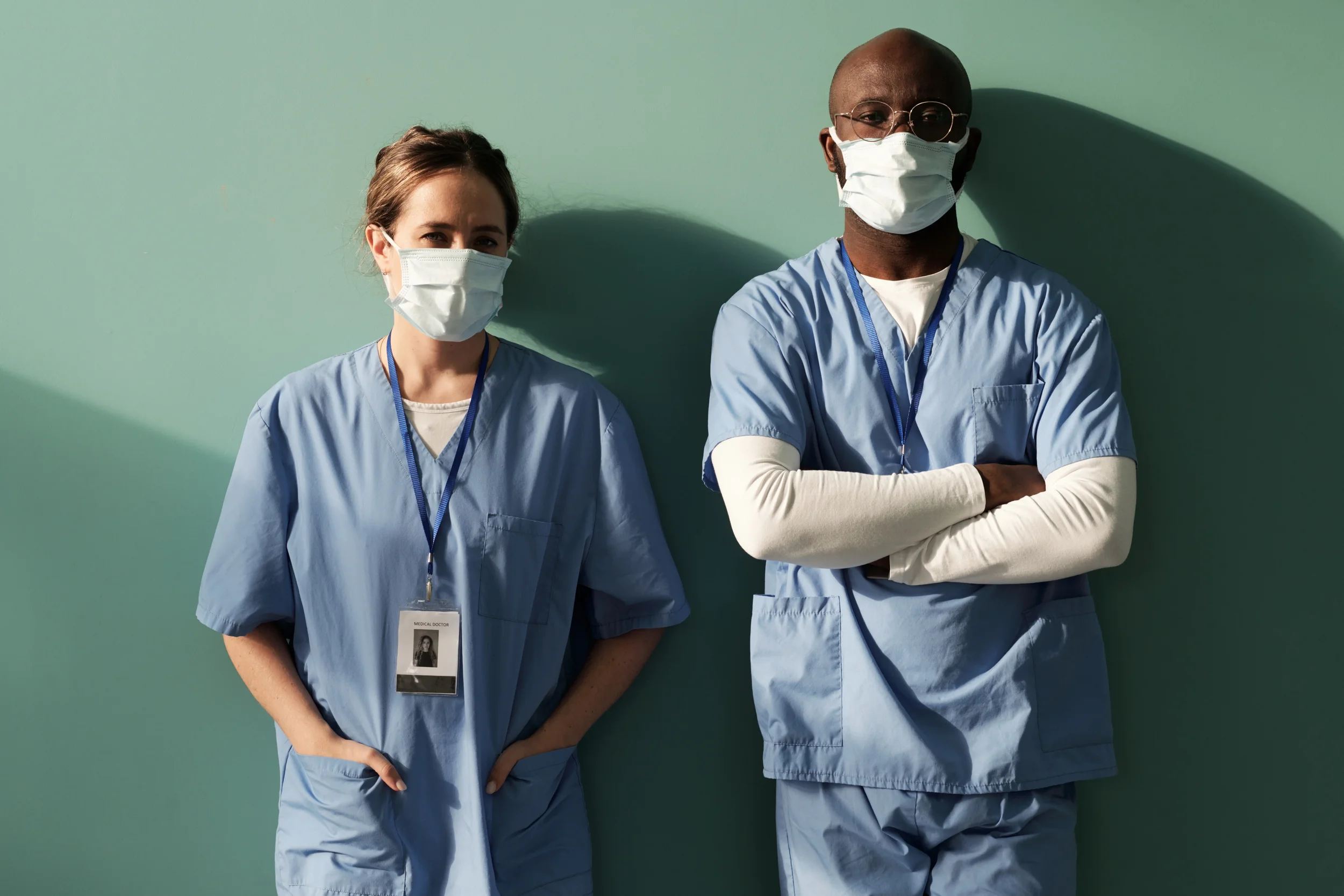 Two healthcare professionals, a woman and a man, standing against a green wall, both wearing light blue scrubs and white face masks. The woman has brown hair tied back, and the man is bald with glasses. The man has his arms crossed, and the woman has a name badge that indicates she is a medical doctor.