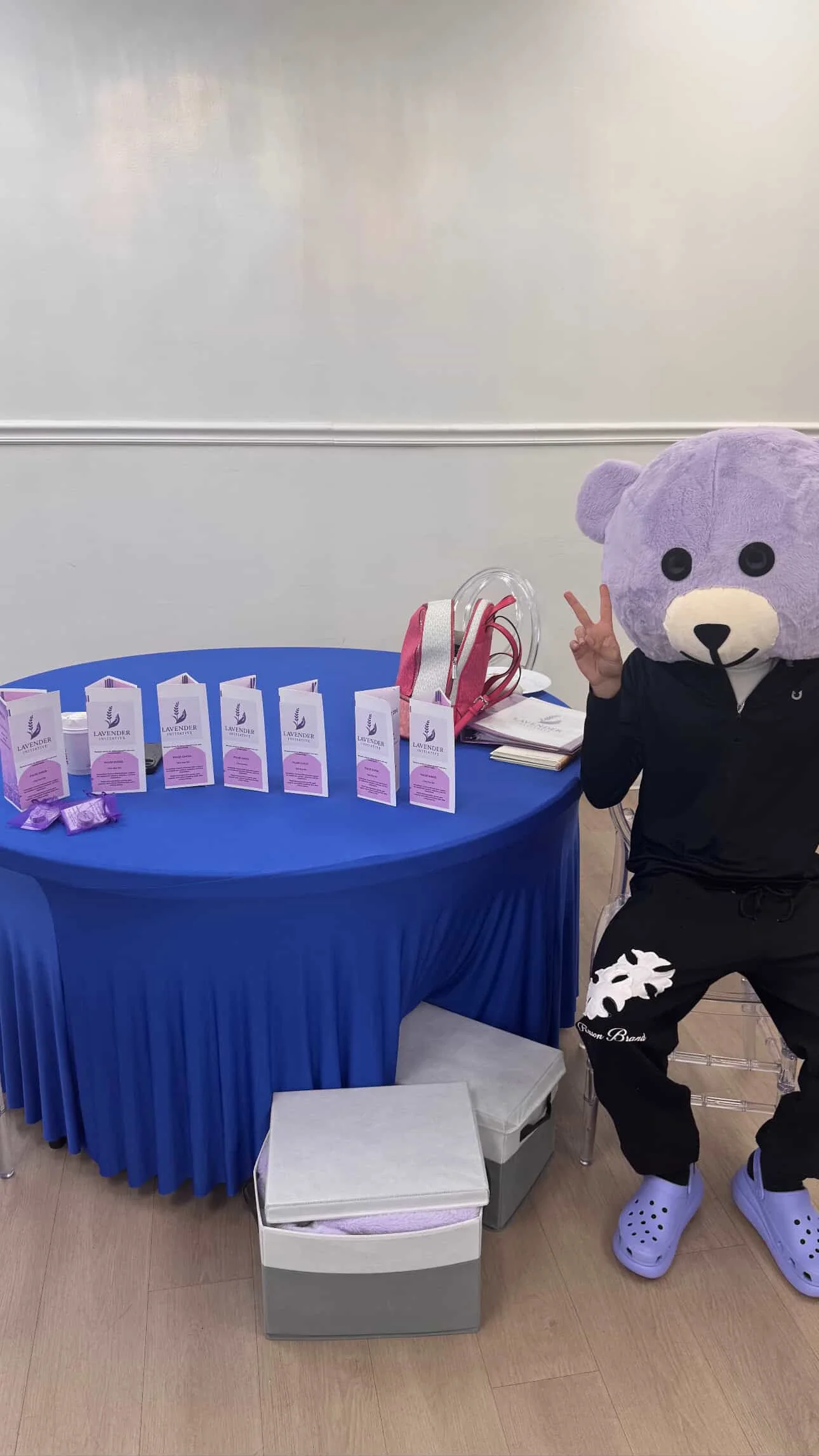 Display table with lavender products, a person in a bear mask and black outfit making a peace sign, seated next to a clear bag, stack of papers, and pink bag.