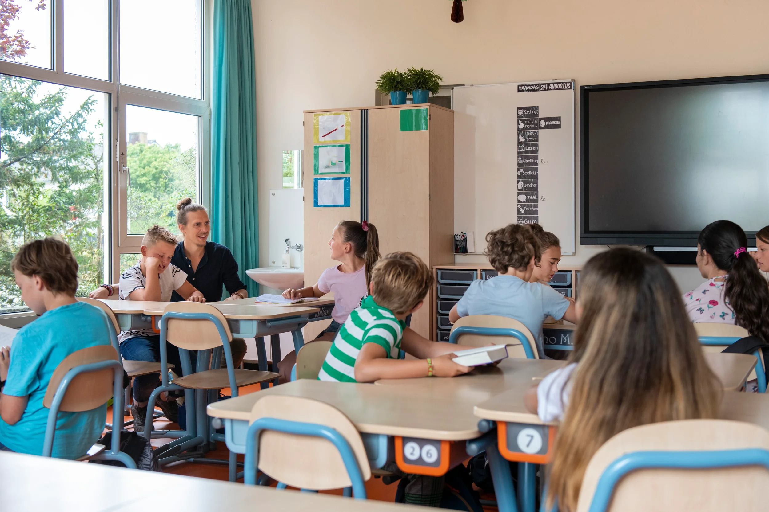 Classroom filled with students sitting at desks, some are reading or talking, with a teacher or facilitator engaging with a student. Large windows with blue curtains let in natural light. There is a blackboard or screen on the wall, and various educational posters are visible.