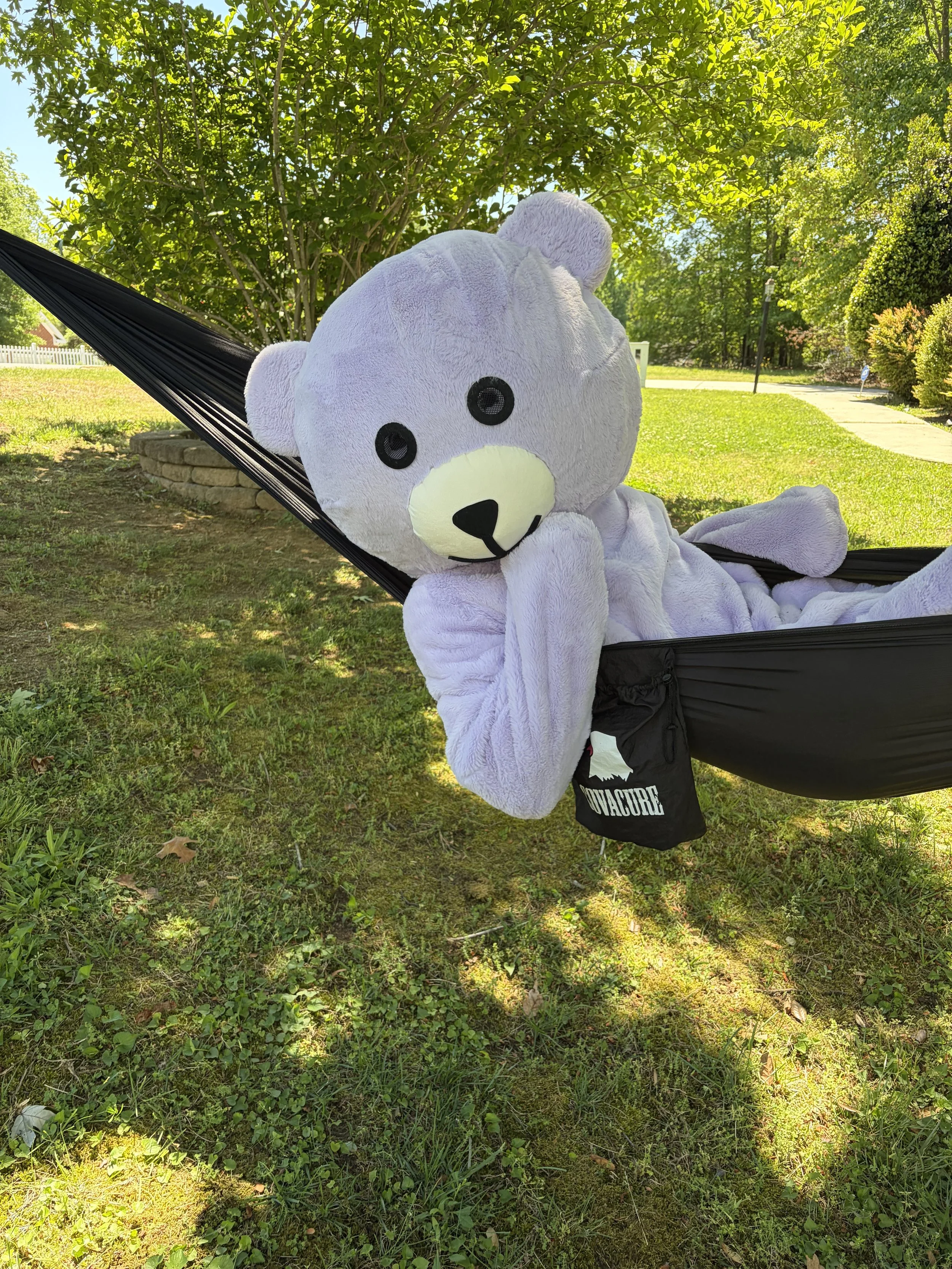 A large white teddy bear with black eyes and a black nose resting in a hammock on a sunny day, surrounded by trees and green grass.
