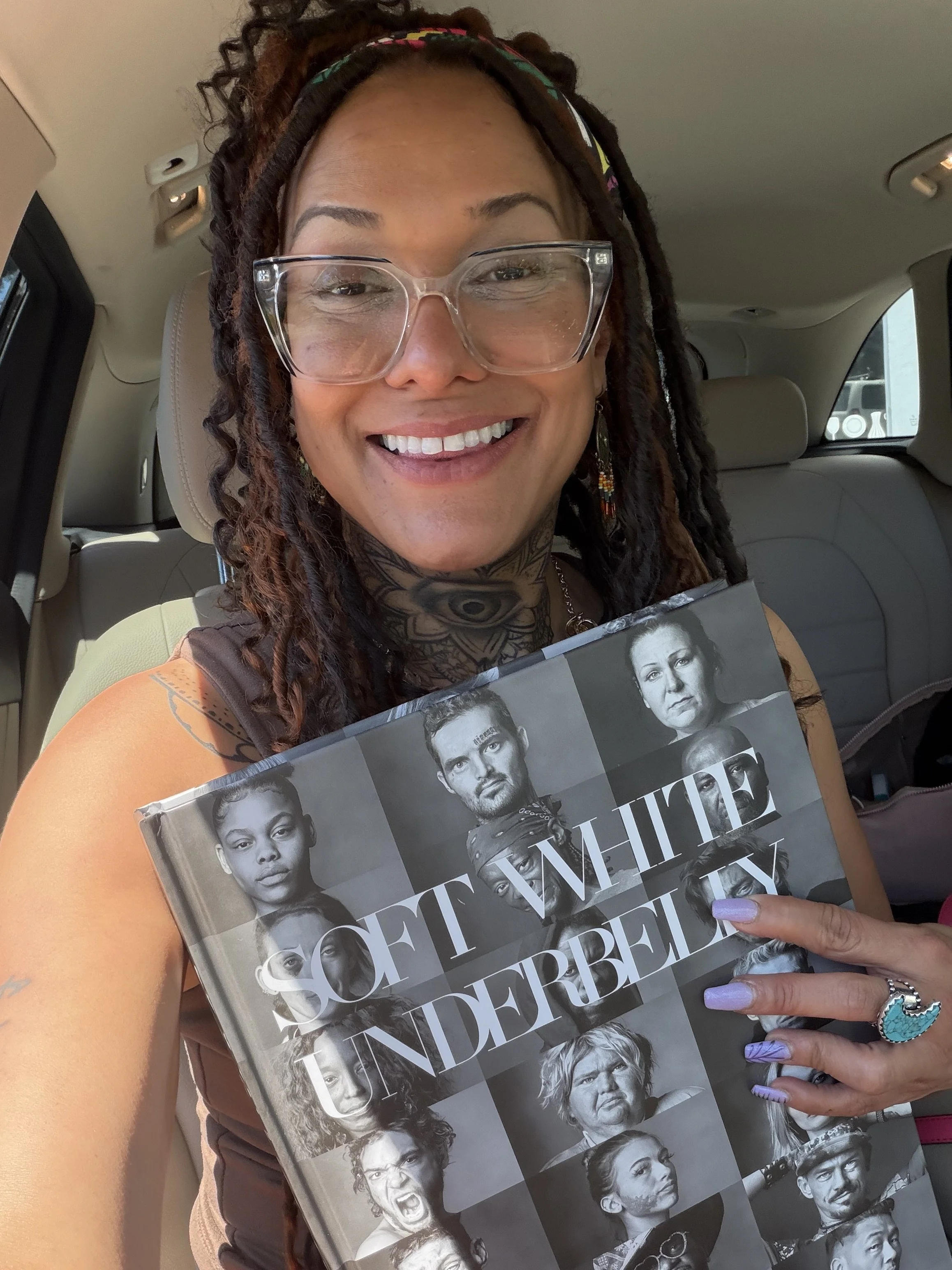 Lavender Initiative founder with goggles and a colorful hair band sitting in car, holding a magazine with black-and-white portraits of various people on the cover.