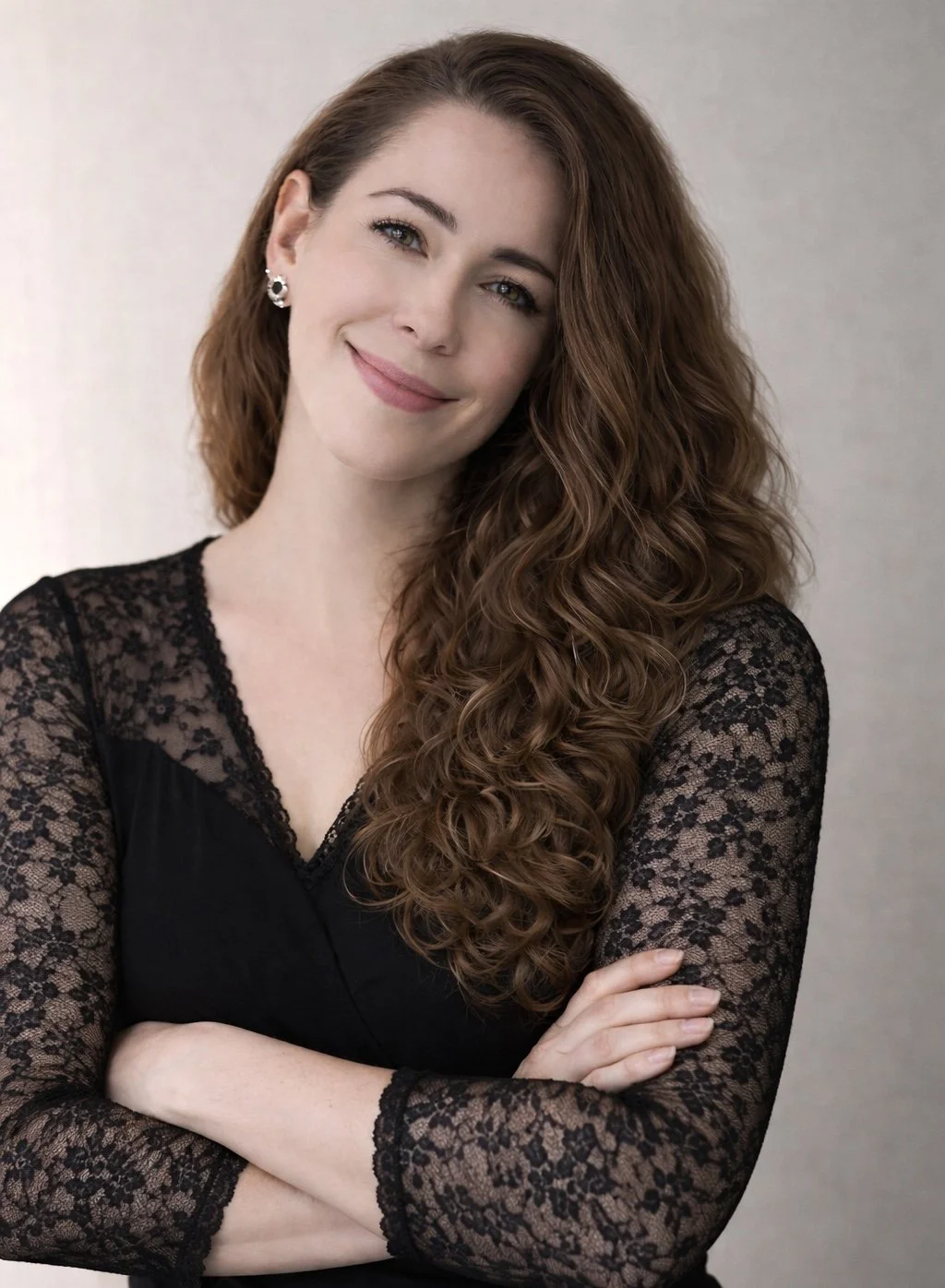 A woman with long, curly, auburn hair smiling with arms crossed, wearing a black lace top and earrings, standing against a plain light-colored background.