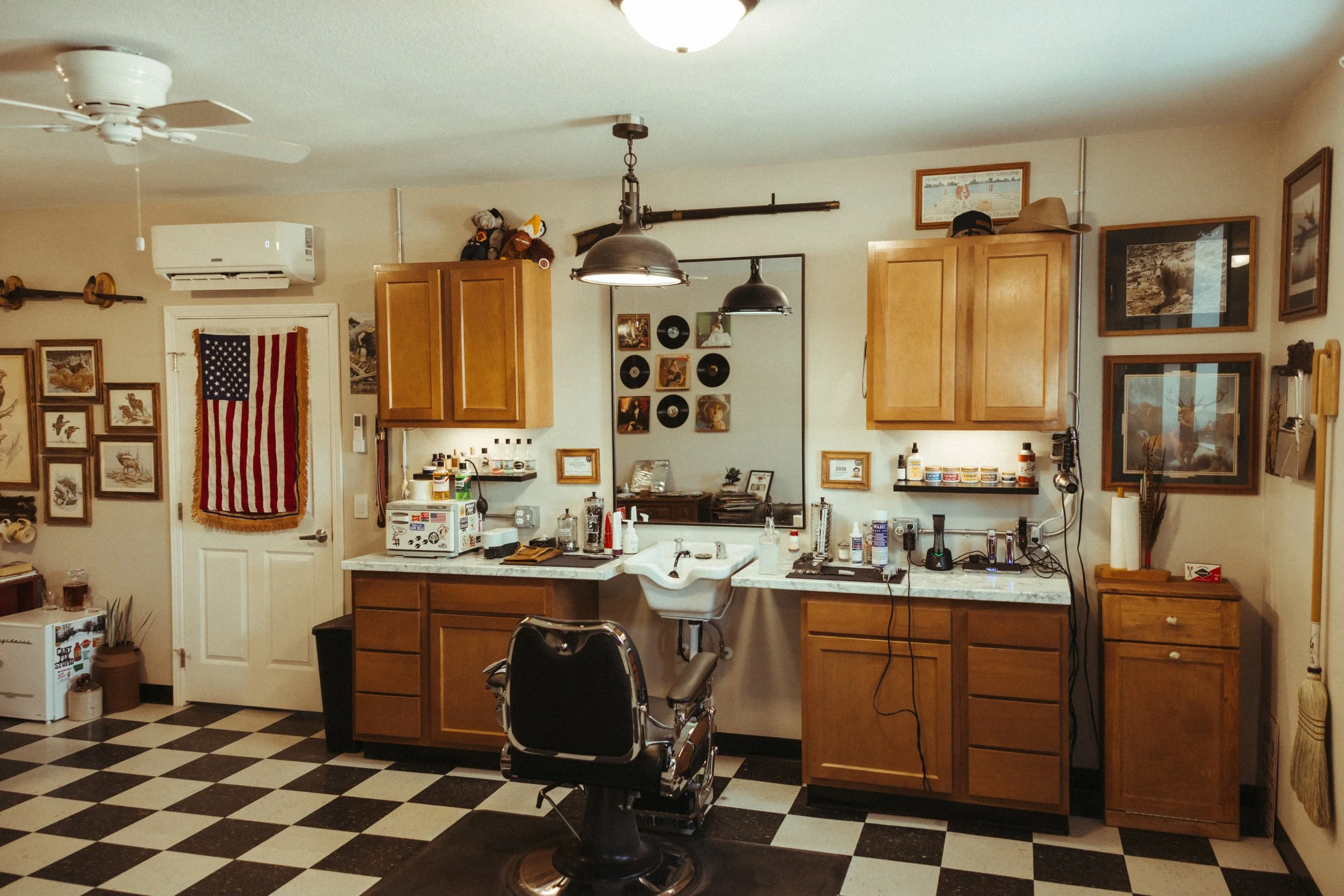 A kitchen with wooden cabinets, a checkered black and white floor, framed pictures on the walls, and a counter with various items including a coffee maker and a small refrigerator. There are ceiling lights, a ceiling fan, and an American flag hanging