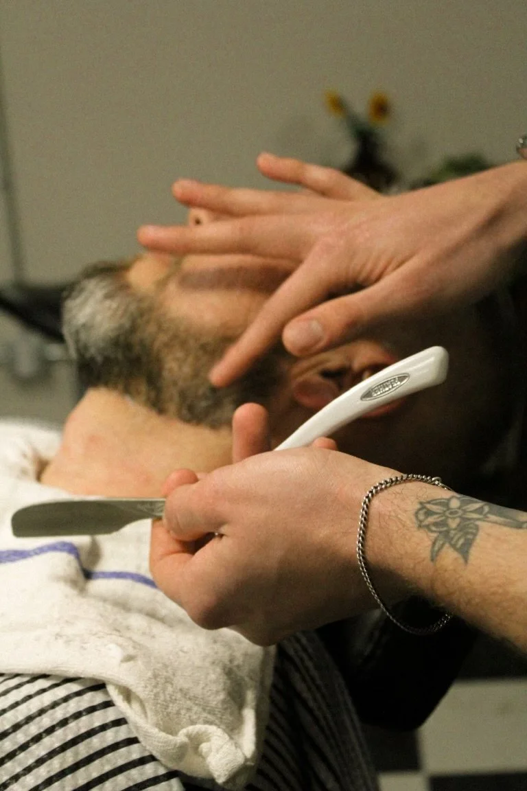A person holding a haircutting razor with a tattooed wrist, trimming a man's beard. The man has red and gray facial hair and is wearing a striped shirt, with a towel draped over his shoulder. In the background, a blurred plant with yellow flowers is visible.