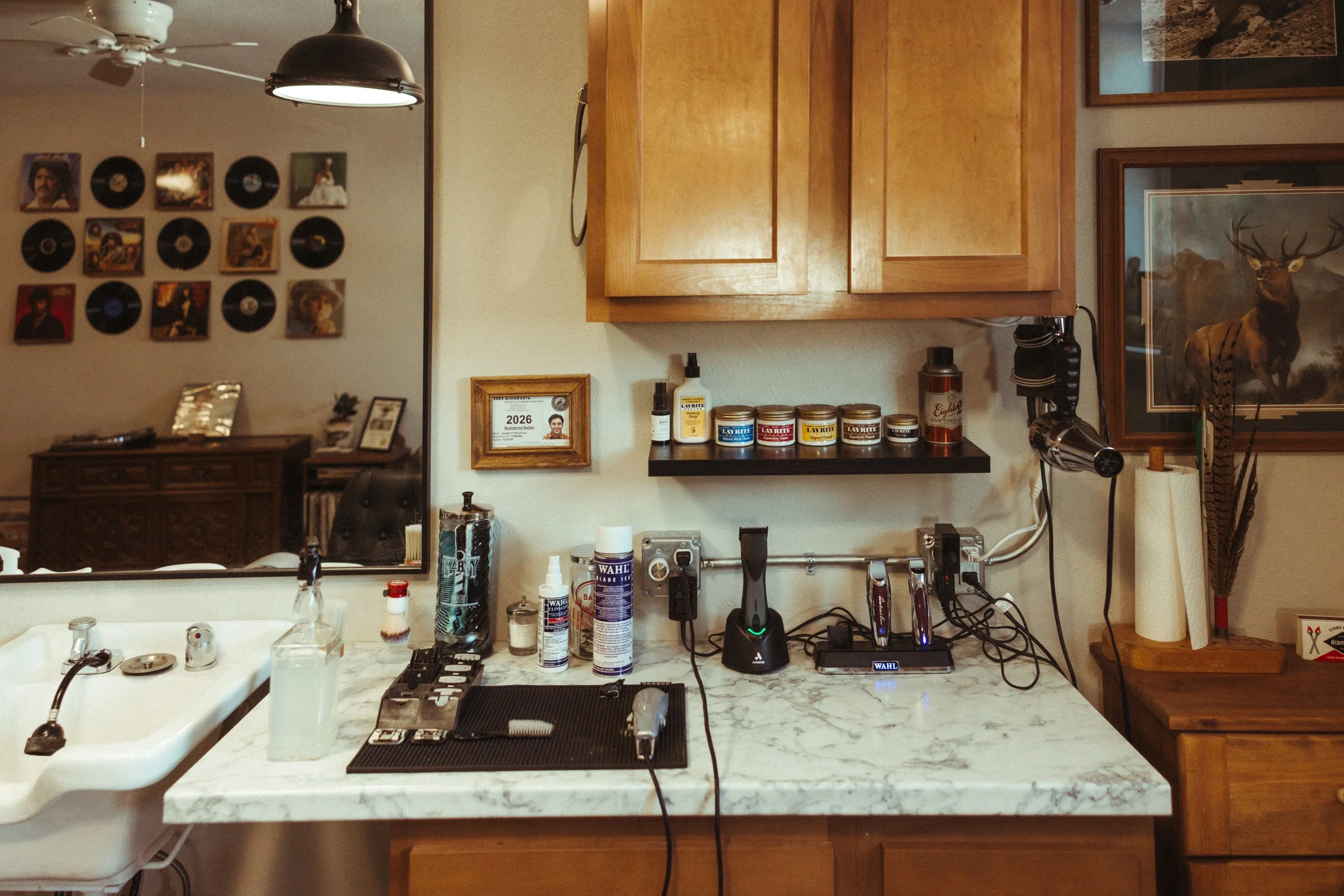 View of a kitchen counter with various grooming and hair products, hair tools, and personal items, with a mirror reflecting a wall decorated with vinyl records and framed photographs, and a wooden cabinet and wall art in the background.