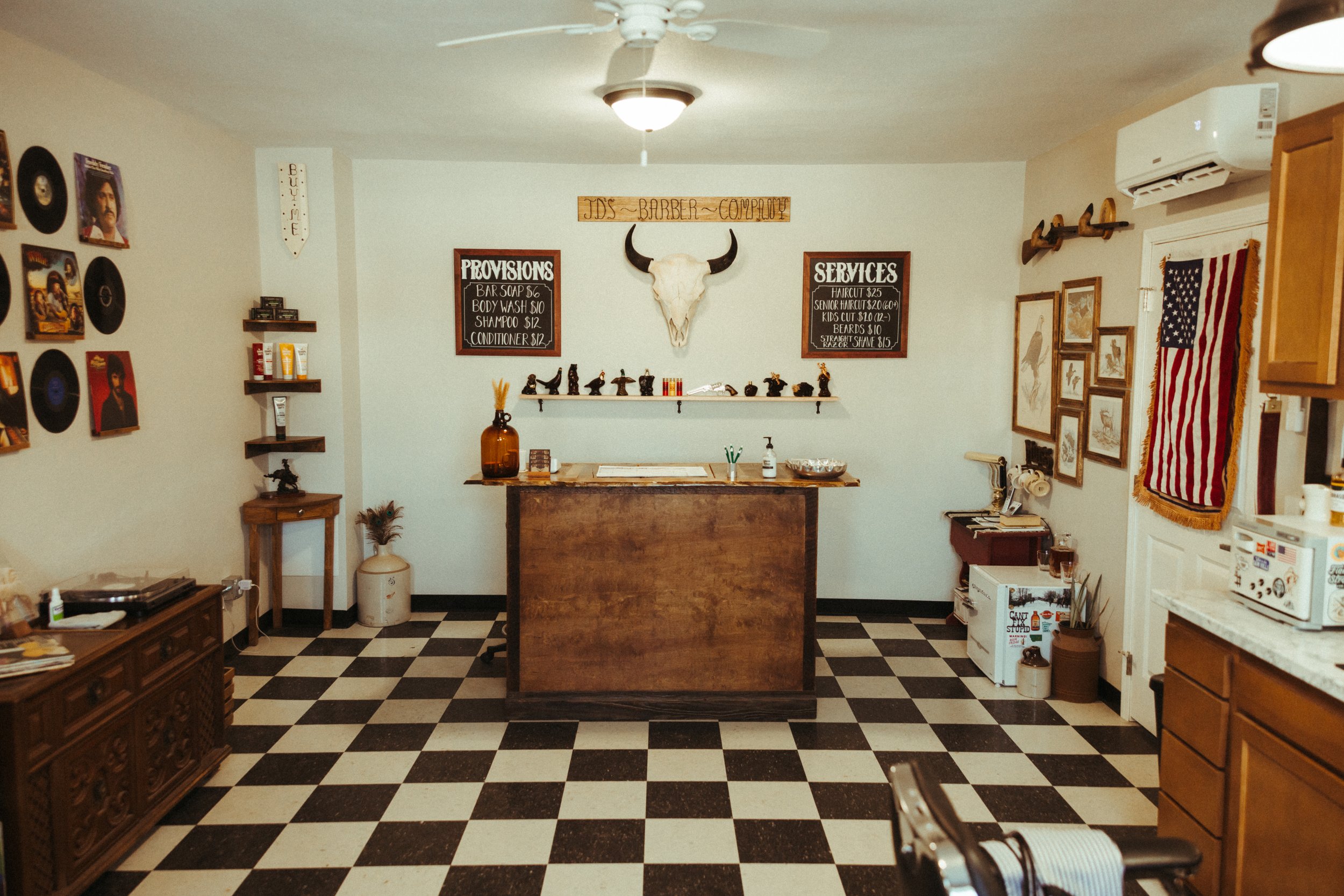 Interior of a barbershop with black and white checkered floor, wooden furniture, framed pictures, wall-mounted vinyl records, a cow skull with horns, and signs detailing services and prices.