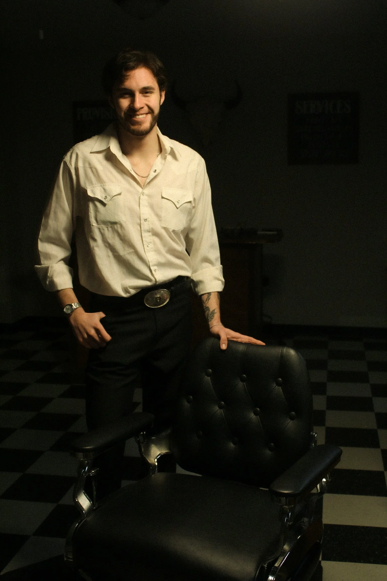 A man with dark hair and a beard, wearing a cream-colored shirt and black pants, standing in a dimly lit room with black and white checkered flooring and black leather chairs.