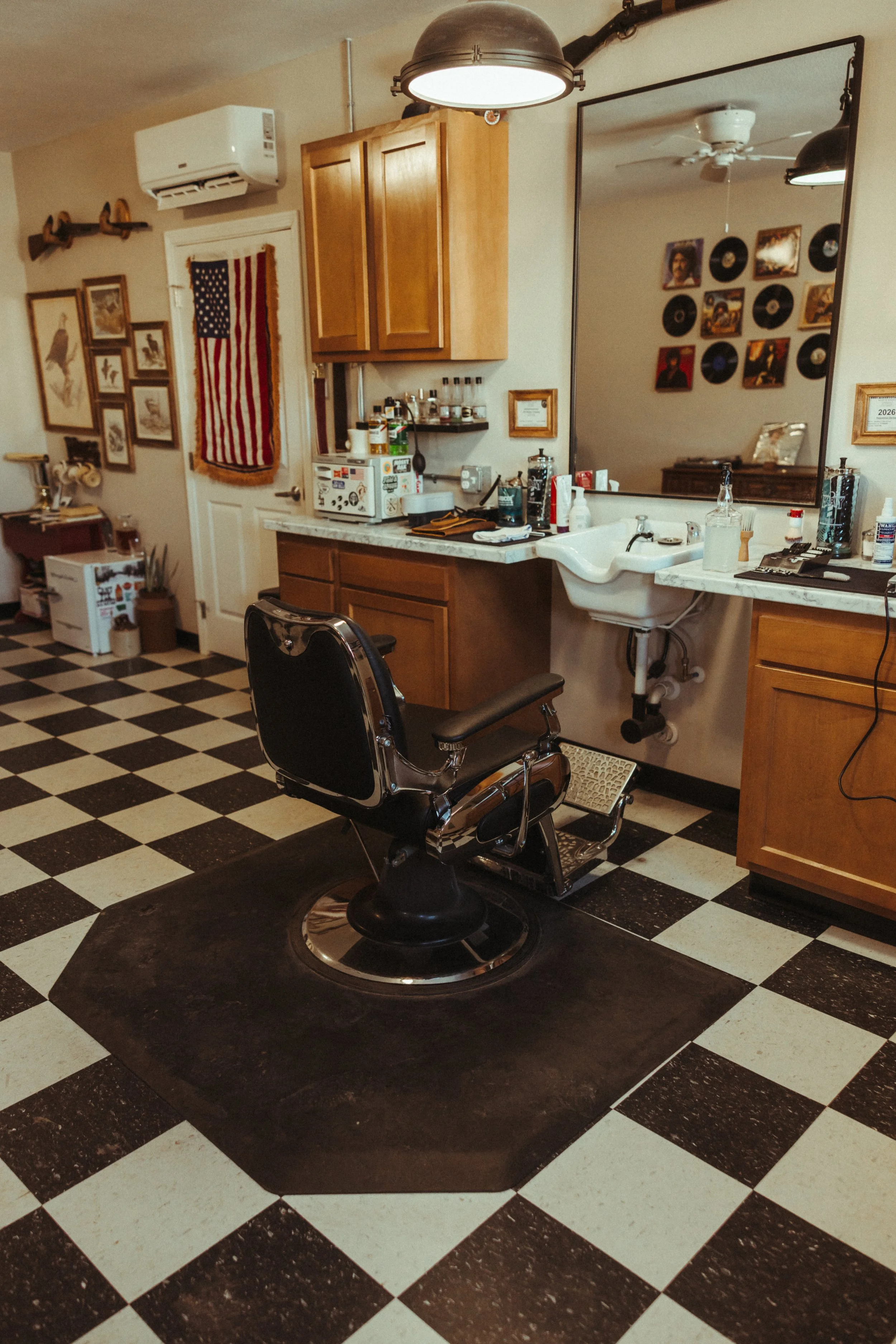 A vintage-style barbershop chair on a black mat in a checkered black-and-white tile floor, with a barber sink, wooden cabinets, a large mirror, and various decor including framed pictures and records on the wall.