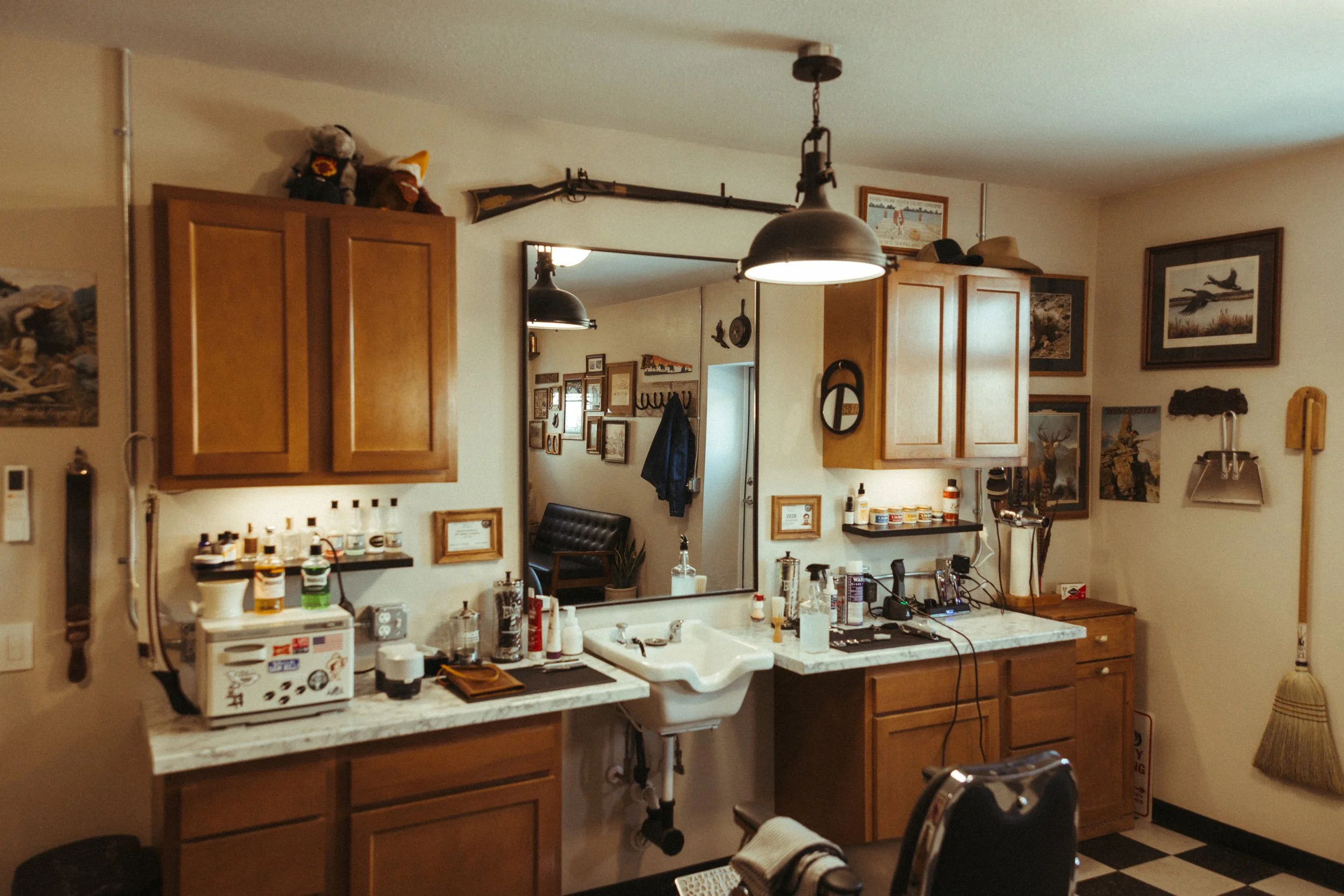 A cluttered kitchen with wooden cabinets, a white countertop, and various items including medicine bottles, electrical cords, and decor. A mirror is mounted on the wall, reflecting another part of the room decorated with framed pictures and a wall ho
