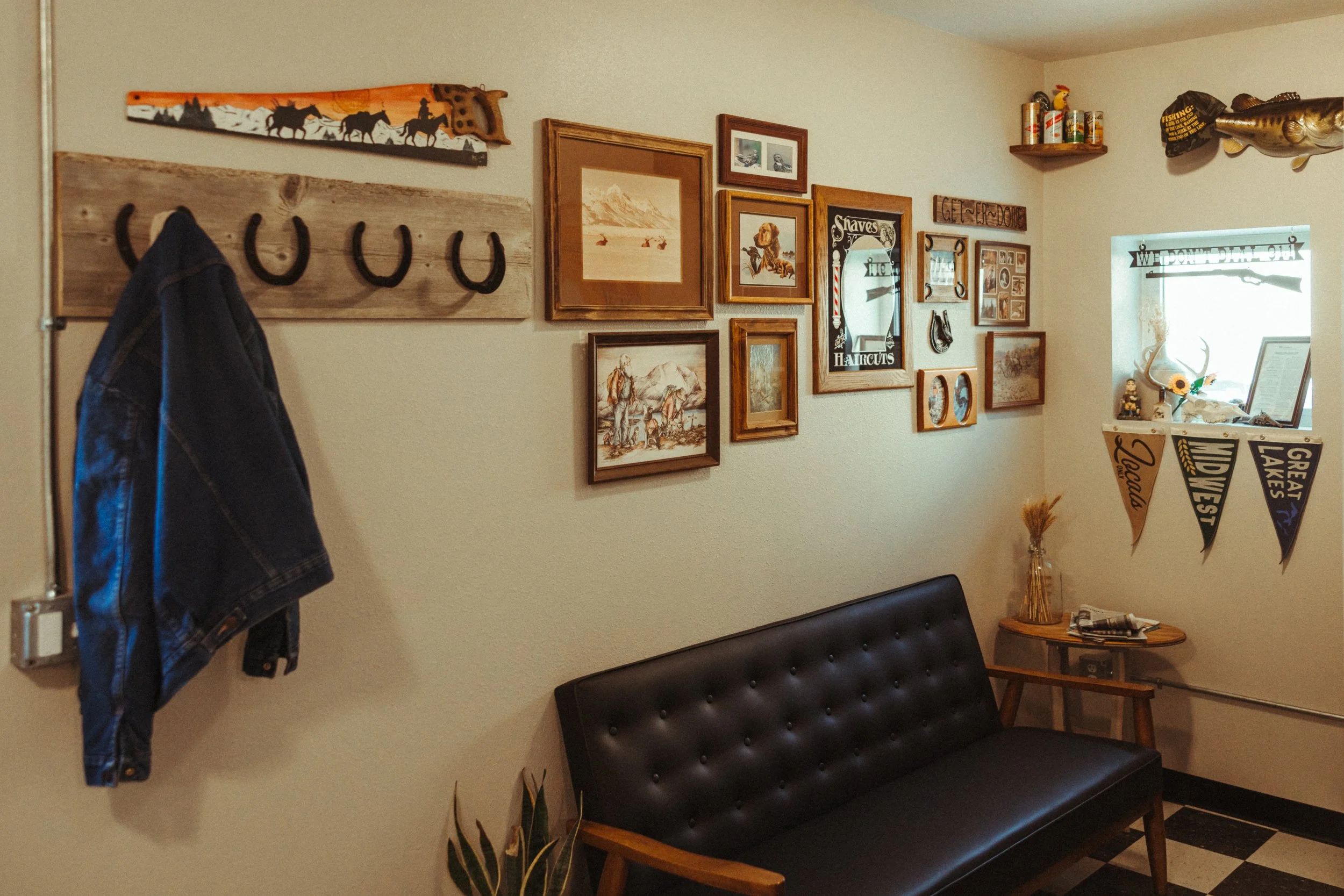 Interior of a living room with a gallery wall, a black leather bench, a small wooden side table, and a window with decorative items, including a fish and fishing-themed decor.