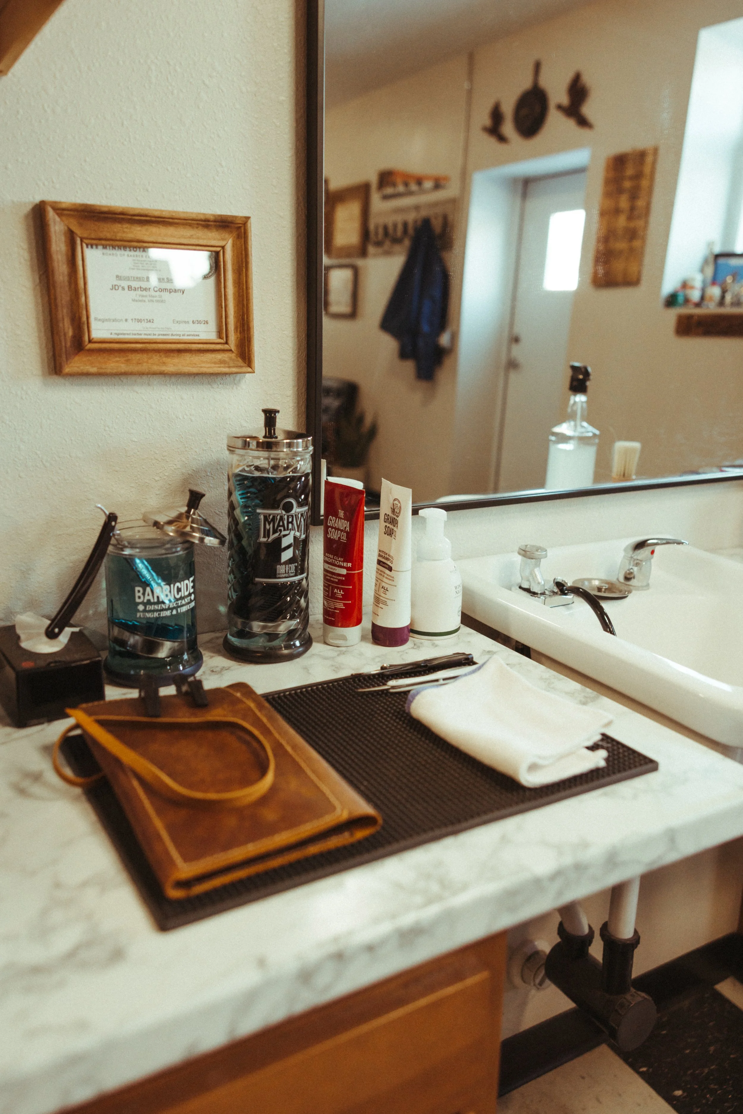 A bathroom vanity with a marble countertop, featuring a white towel, a leather case, and various toiletries including creams and sprays. A sink with a chrome faucet is visible, and a large mirror extends across the countertop. On the wall above the m