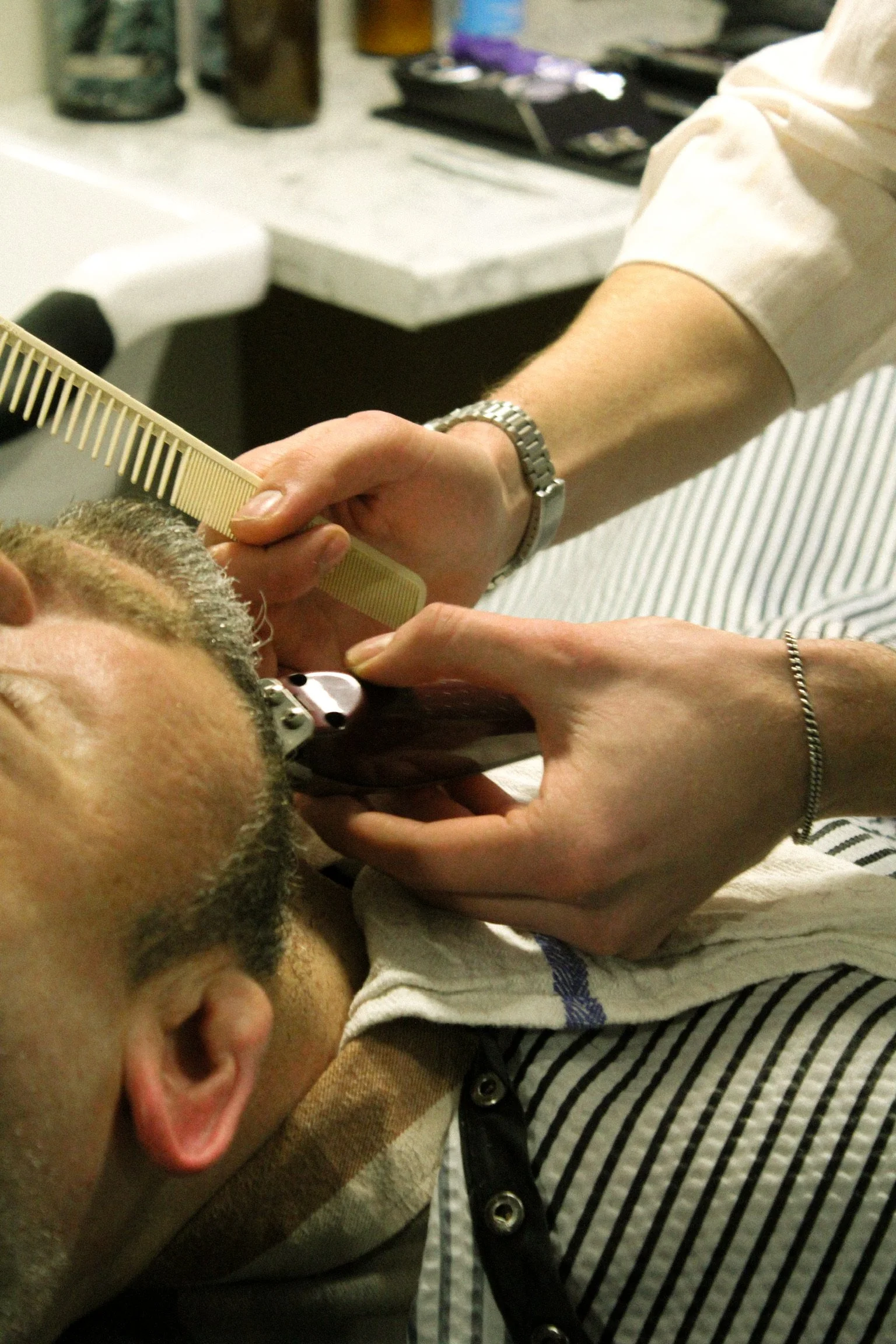 A person being shaved by another person using an electric device, with a comb seen in the hand of the person doing the shaving.