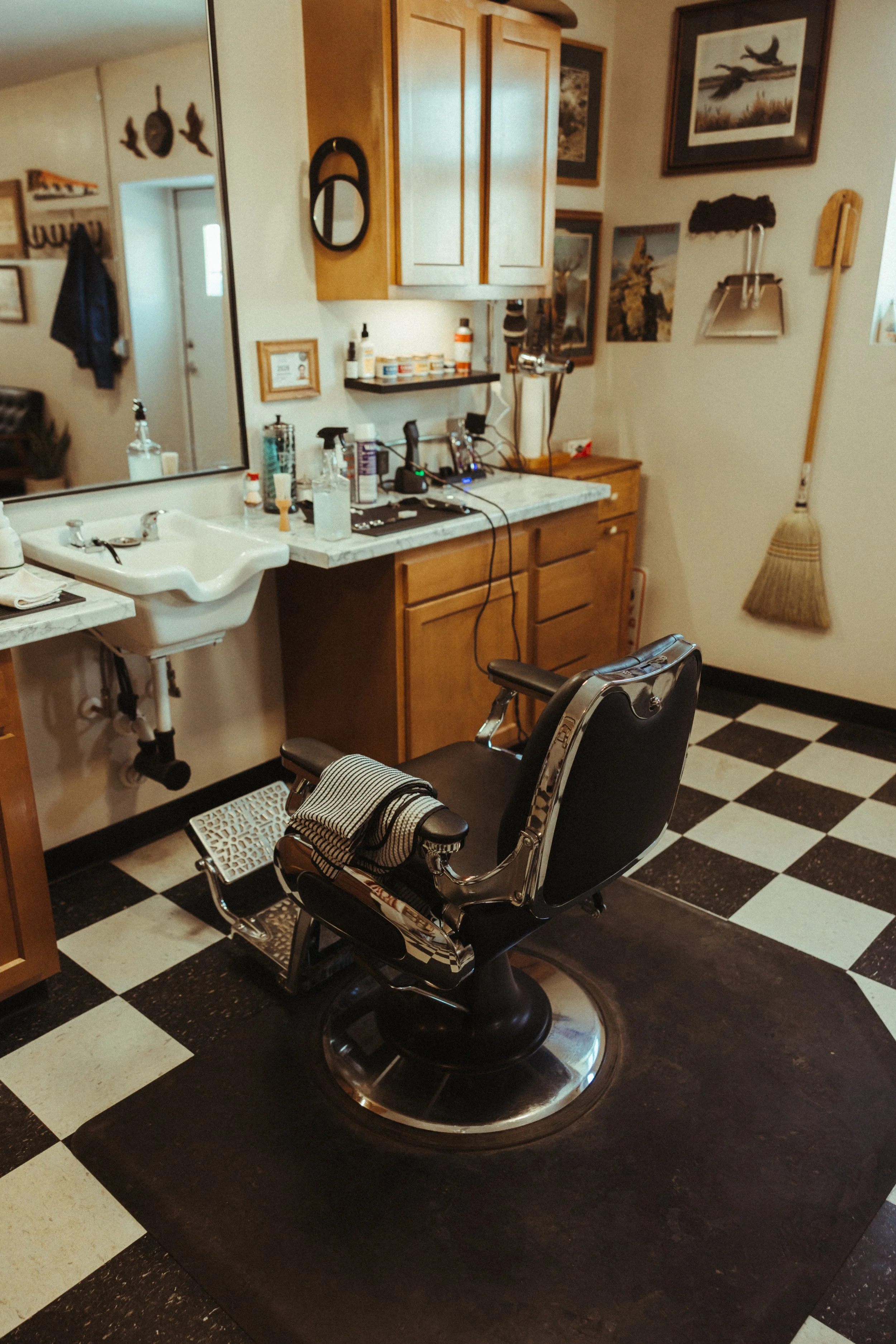 Image of a barber shop chair in front of a mirror and a sink, with walls decorated with framed pictures, a broom, and various tools on the counter.