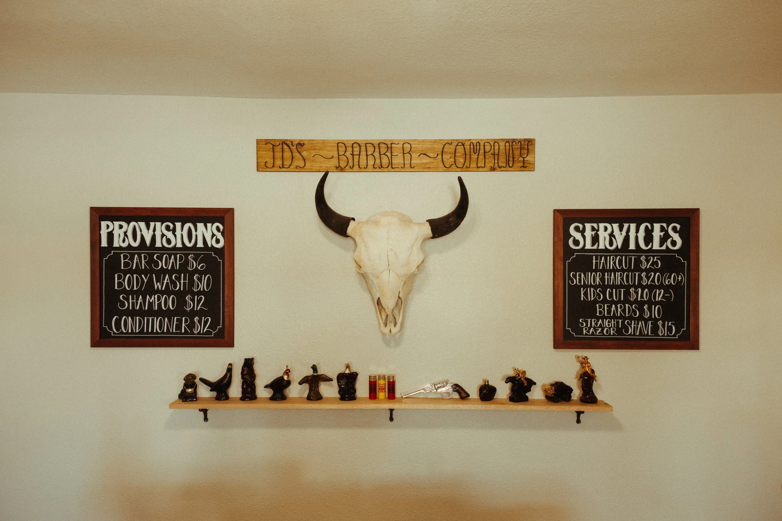 Interior of a barbershop with a mounted cow skull with horns on the wall. On the wall, there are two framed chalkboards listing grooming services and prices, and a wooden sign above the skull reads "TDS - BARBER - COMPANY." A shelf below the signs di