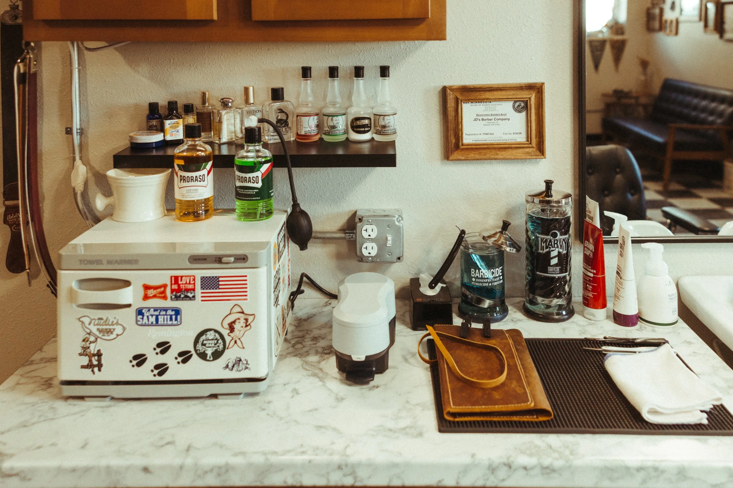 A countertop with various bottles of aftershave, skincare, and grooming products, a small white appliance with stickers, a cup, a black electric shaver, and a glass of water. A framed certificate is hanging on the wall. The background shows part of a
