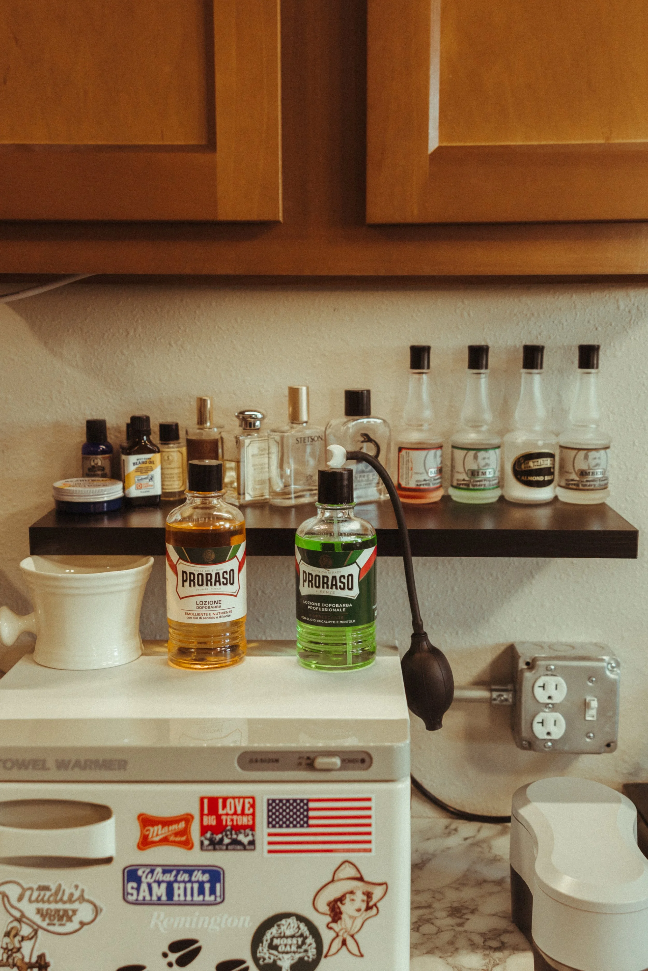 A counter with various bottles, including Proraso aftershave, a mug, and a small container. A microwave oven is partially visible with stickers, and a power strip with multiple plugged-in appliances is behind it. Wooden cabinets are above the counter.