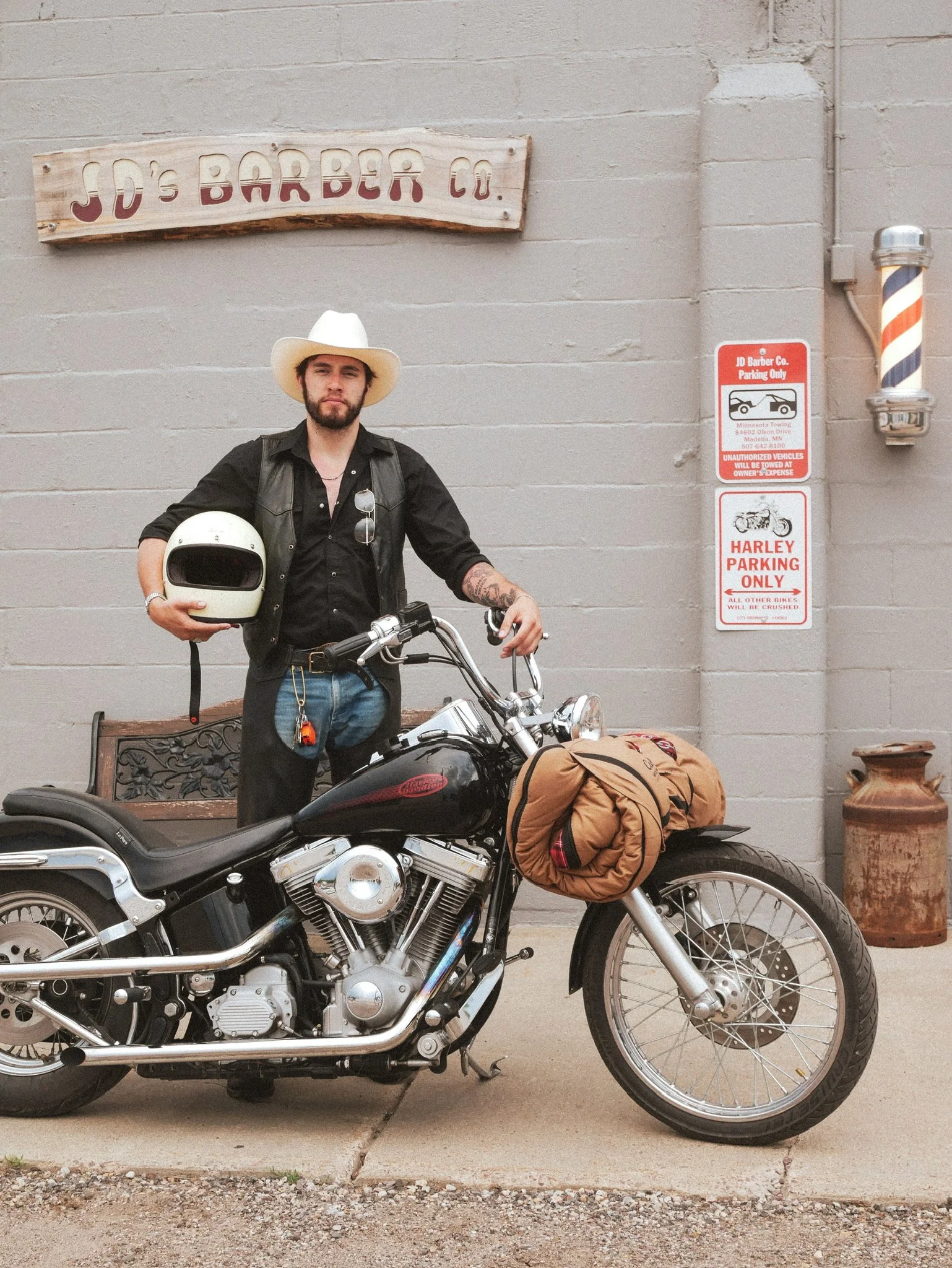 A man with a beard and tattoos, wearing a white cowboy hat and black shirt, standing next to a black motorcycle. He is holding a white helmet and standing in front of a beige brick wall with signs for JD Barber Co. and Harley parking only. There is a wooden bench and a large ceramic pot nearby.