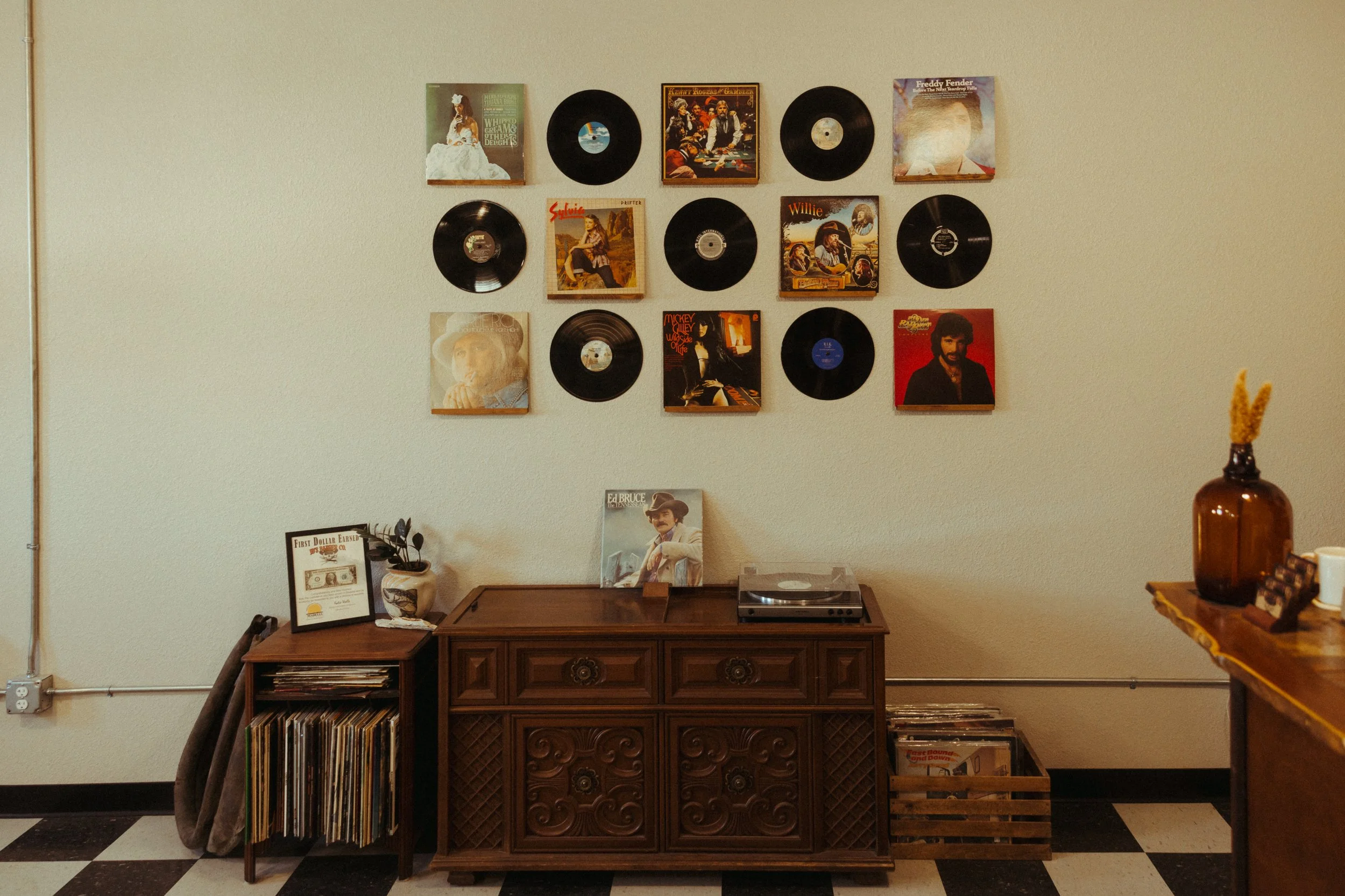 A vintage wooden sideboard with a record player on top, filled with vinyl records and decorative items, against a beige wall decorated with framed vinyl record covers and vinyl records.