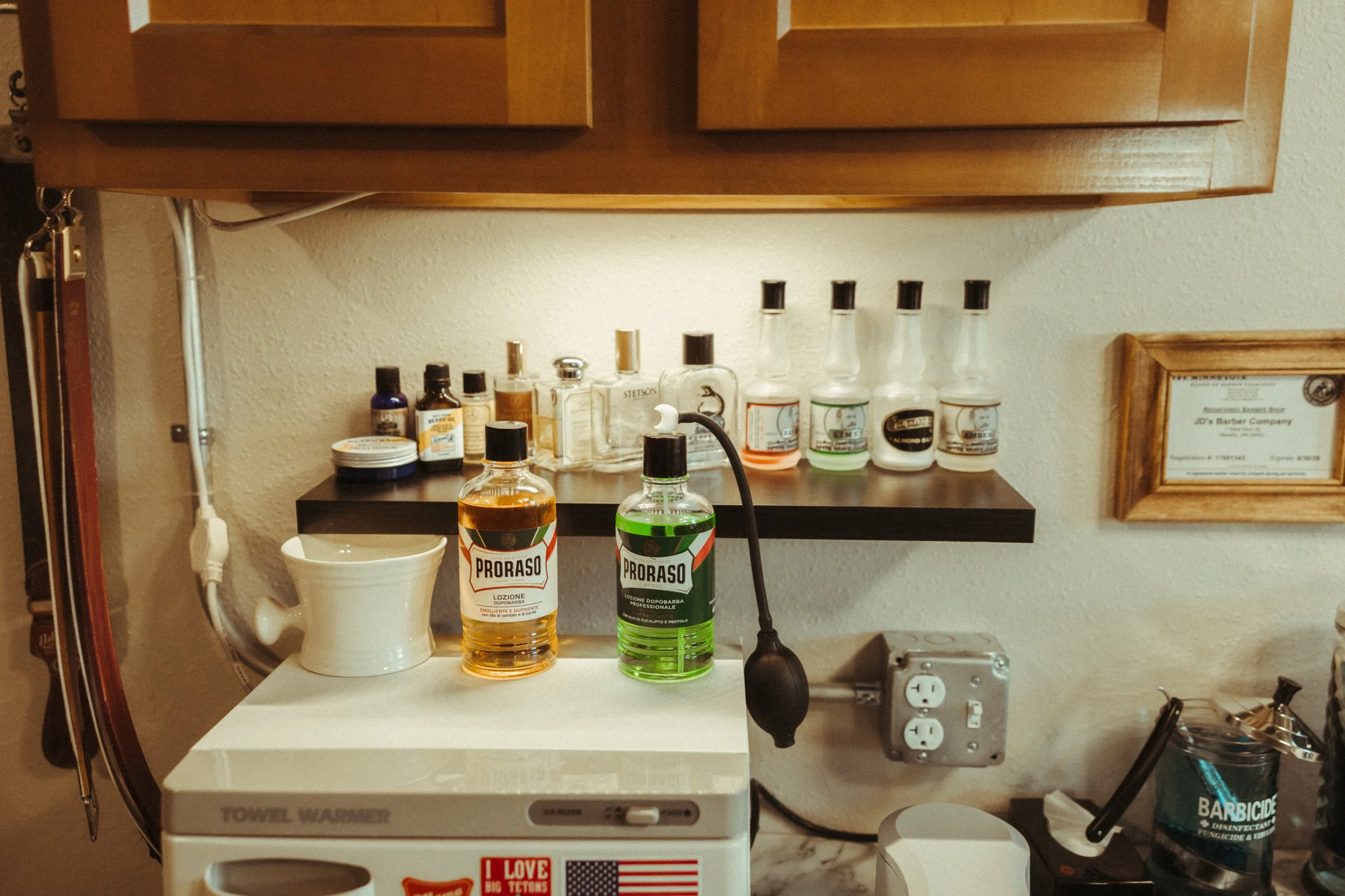 A small black table with various bottles and containers, some labeled Proraso, and a white ceramic mug. Behind the table is a wall with a framed certificate, power outlets, and a shelf with medicine bottles and spray cans. A black cord hangs from abo