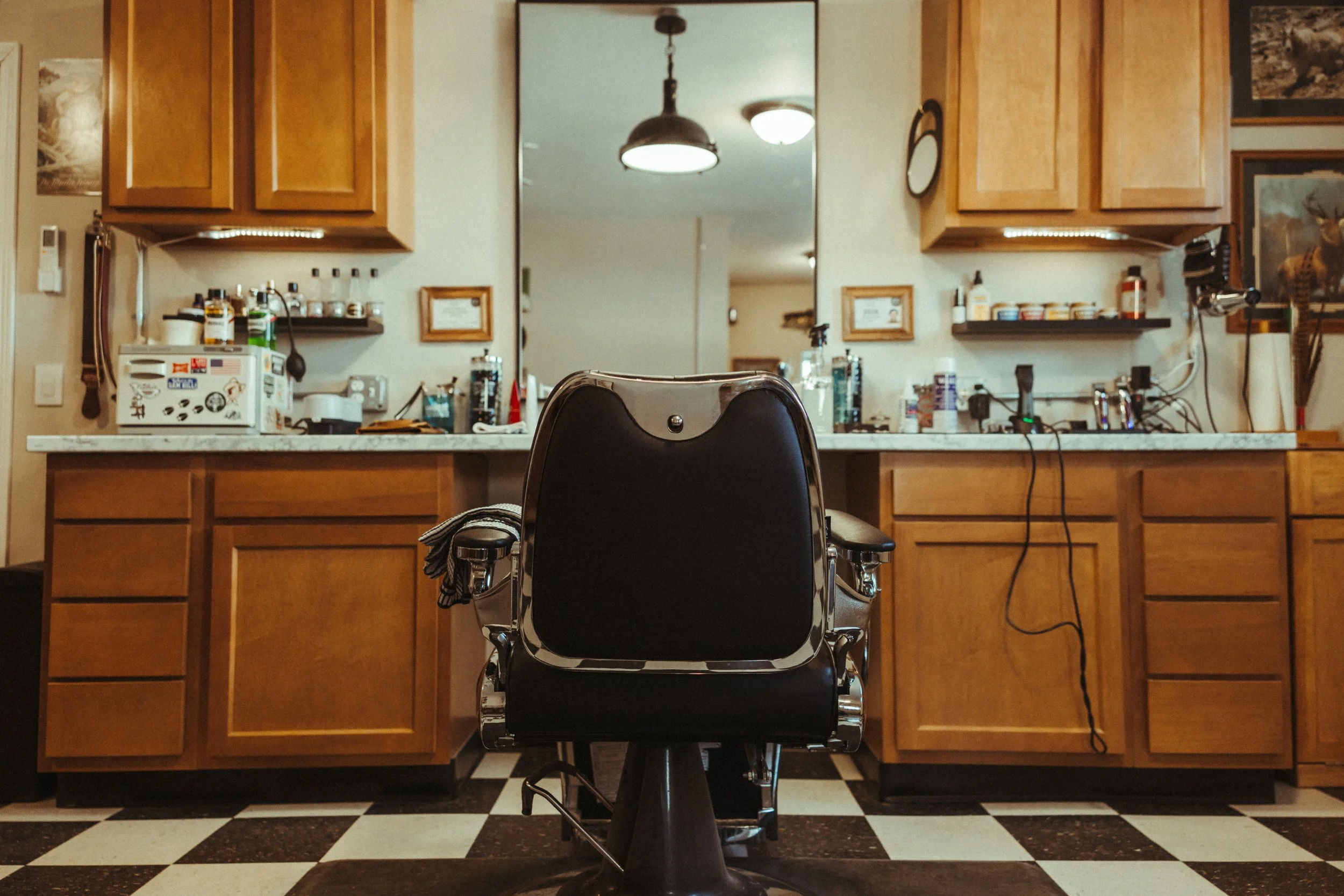 A barber chair facing a large mirror in a salon or barber shop. The background features wooden cabinets, various hair styling products, and a checkered black and white floor.