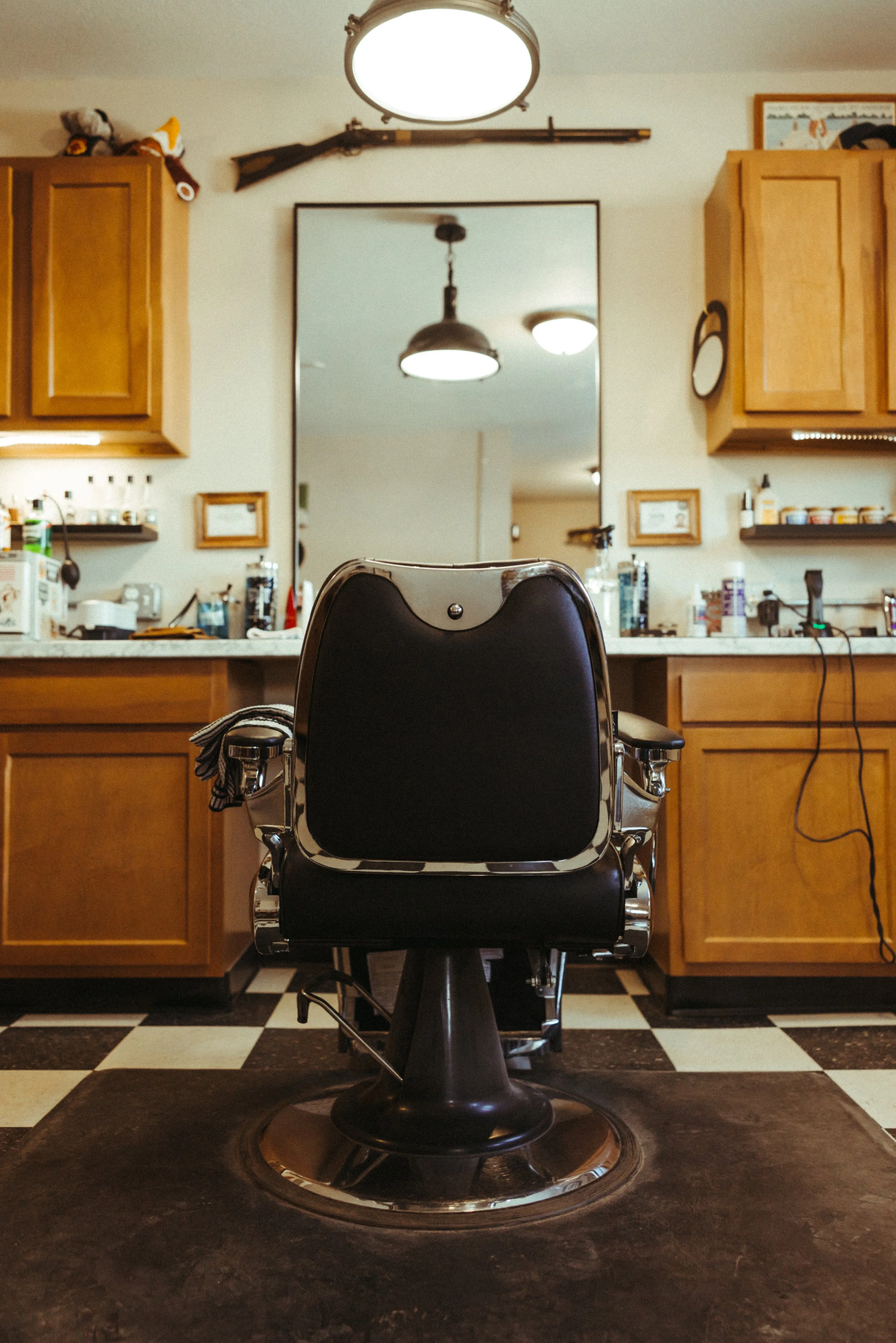 Barber shop chair in front of a mirror with wooden cabinets on the sides, a rifle mounted above the mirror, and a picture frame on the right wall.