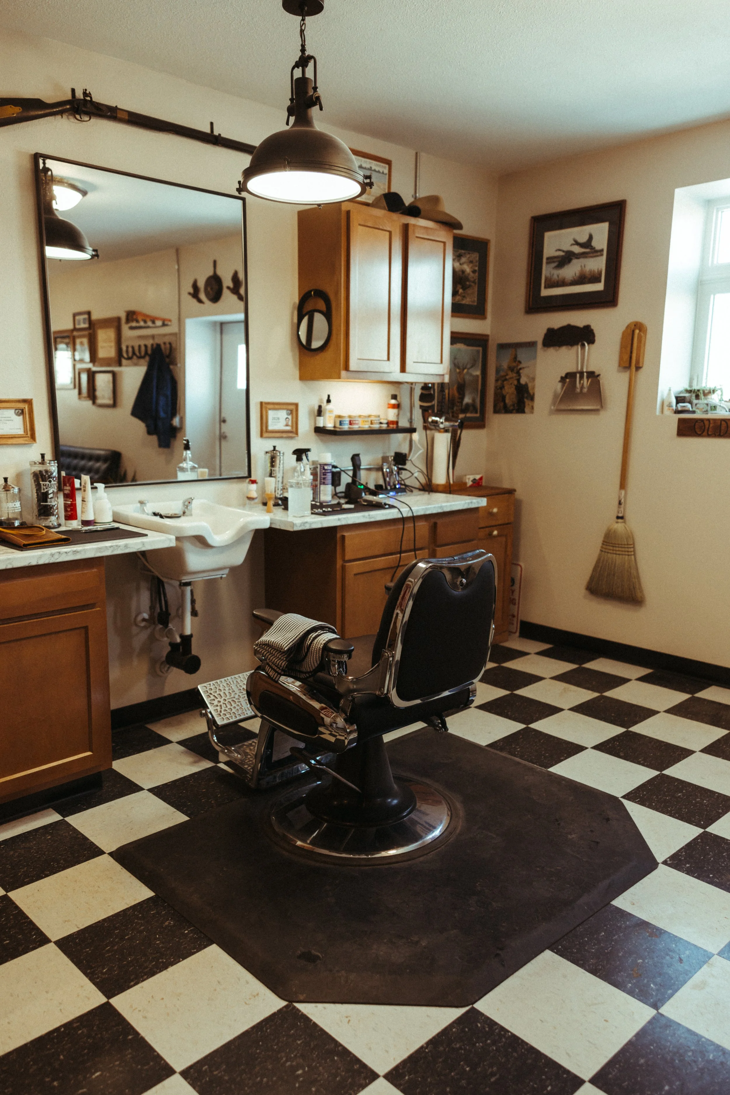 Barber's chair in a vintage-style barber shop with a checkered black and white floor, surrounded by wall art, framed pictures, and a cabinet with hats on top. The chair is black with chrome accents, and there are various barber tools and products on the counter.