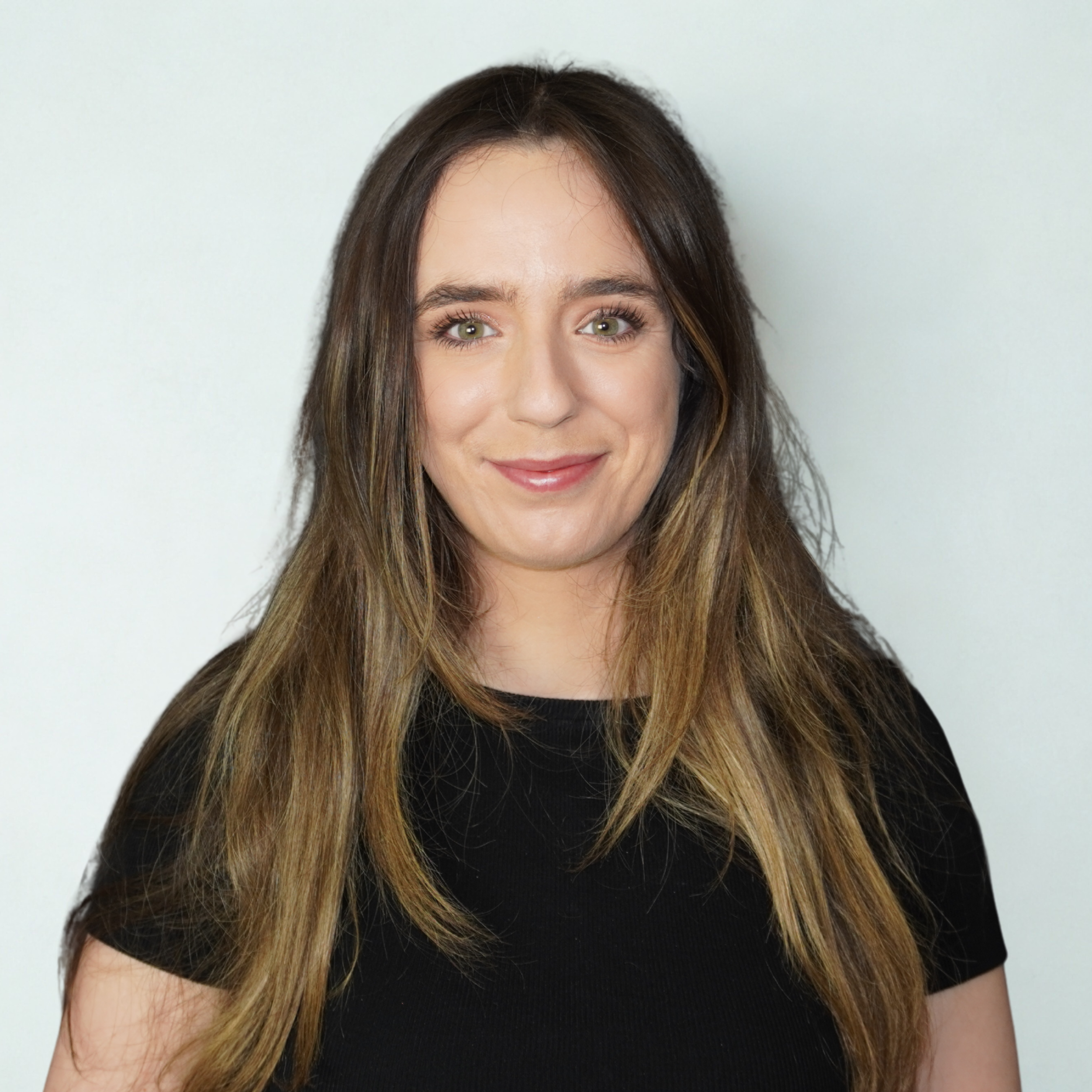 A young woman with long brown hair, green eyes, and a friendly smile, wearing a black shirt, standing against a plain light background.