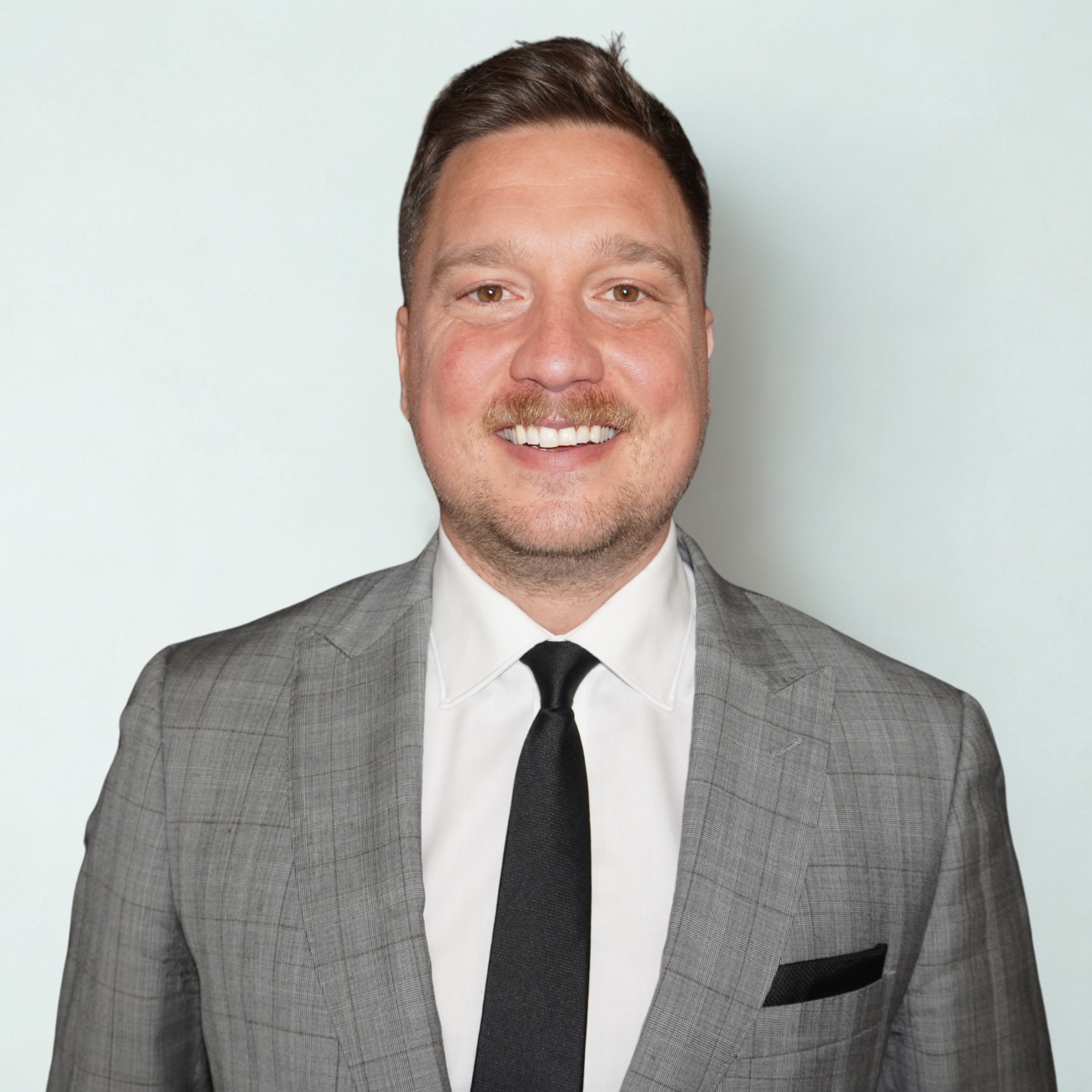 A smiling man wearing a gray plaid suit, white shirt, and black tie in front of a plain light background.